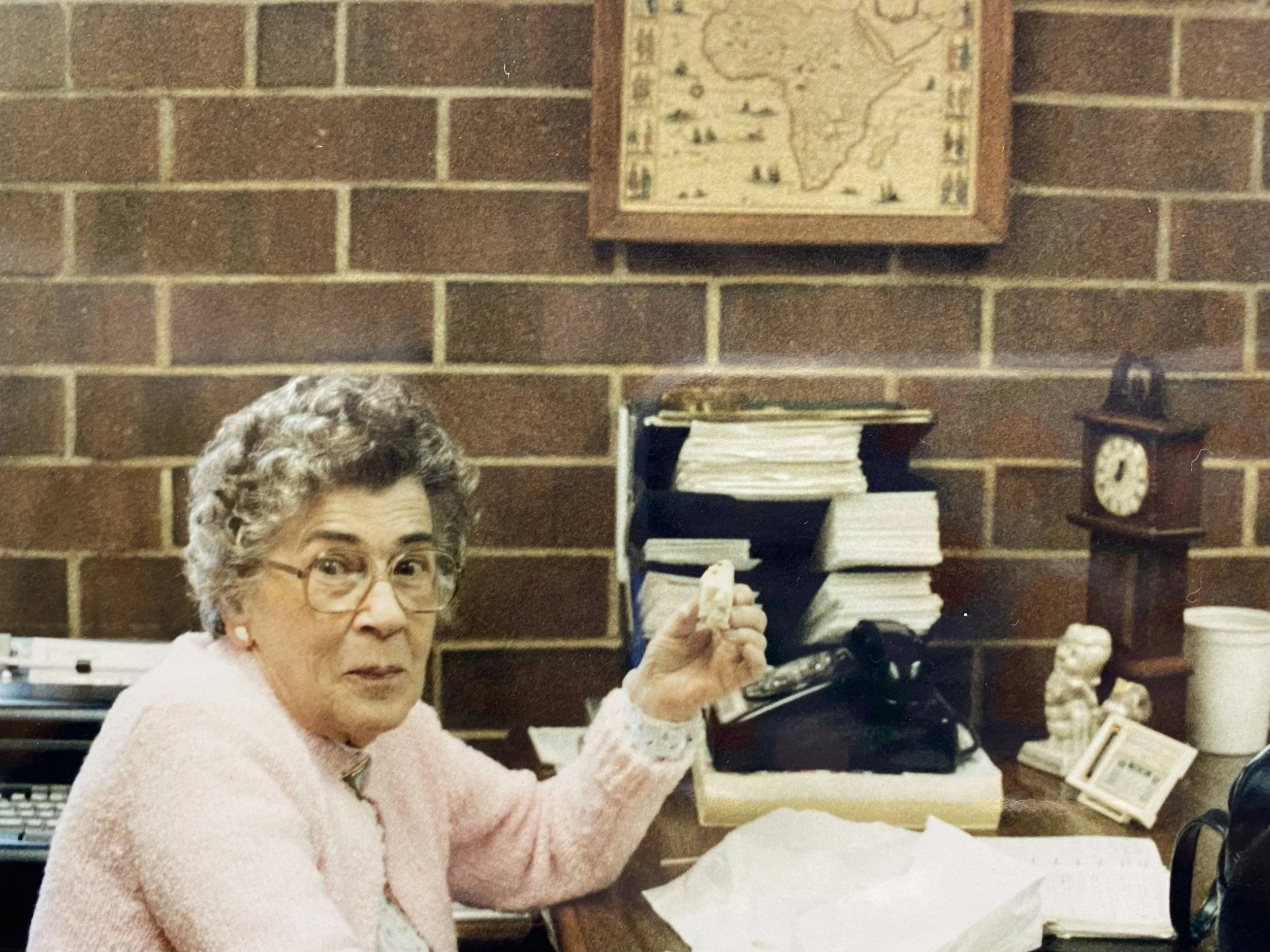 An elderly woman with gray hair and glasses holding an antique telephone, sitting at a desk with stacks of papers and a clock, in a room with brick walls and a framed map.