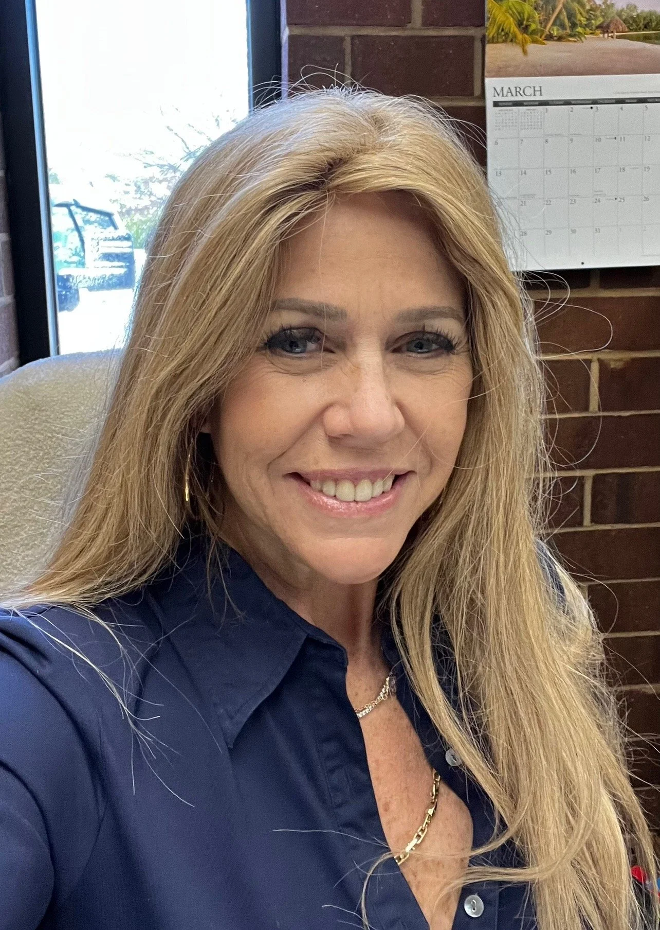 Close-up photo of a smiling businesswoman with blonde hair, wearing a navy blue shirt, gold jewelry, and sitting indoors near a window with a brick wall and a calendar in the background.