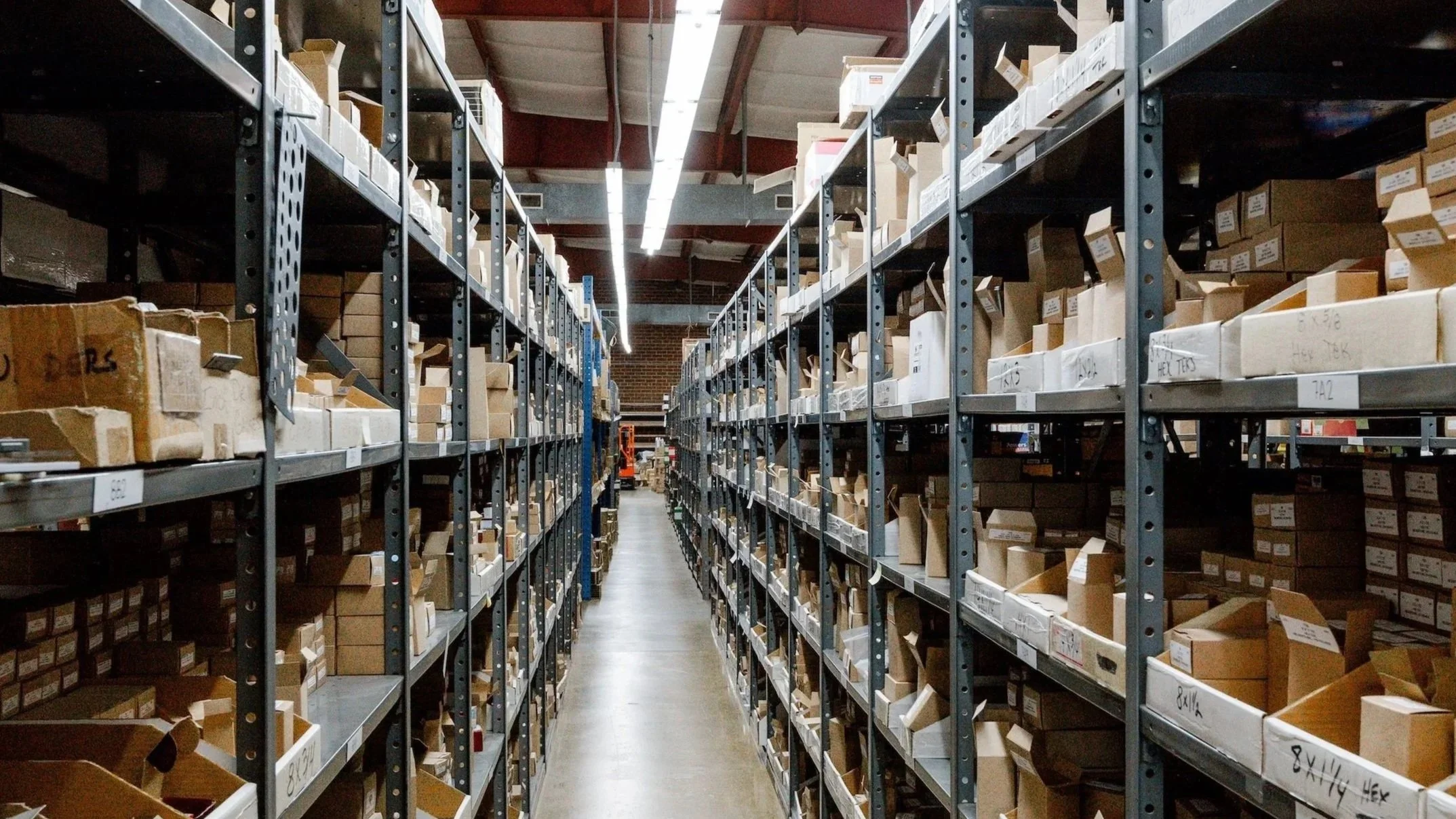 A warehouse aisle with tall metal shelving units filled with boxes of various sizes, some open and some sealed, with handwritten labels on the boxes and shelves. The aisle extends into the background, with a brick wall and ceiling visible at the end.