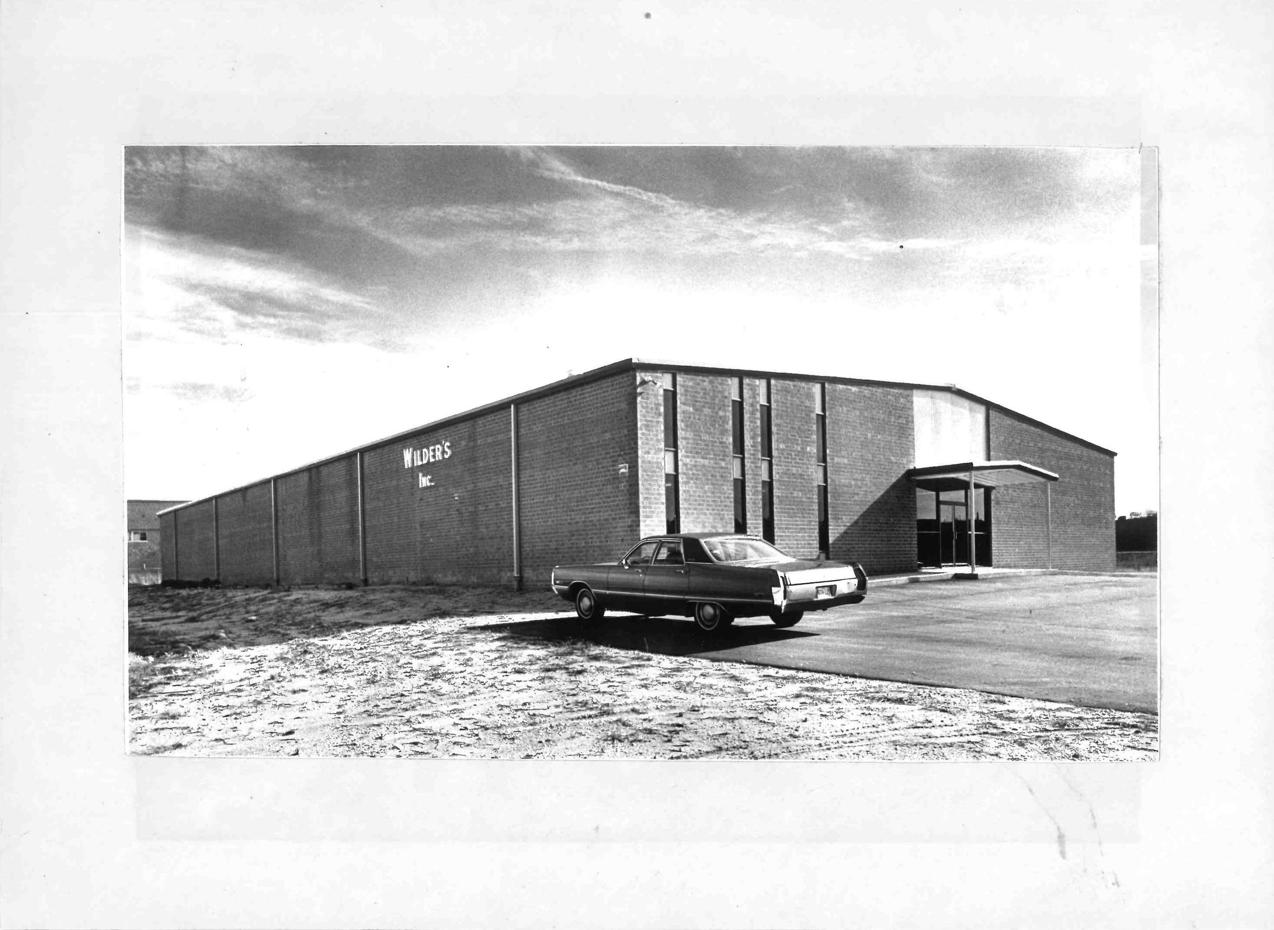 Black and white photograph of a mid-20th century modern brick building with a flat roof and large windows, with a vintage car parked in front. The building has a sign that reads 'Wilder's Inc.' and stands against a partly cloudy sky.