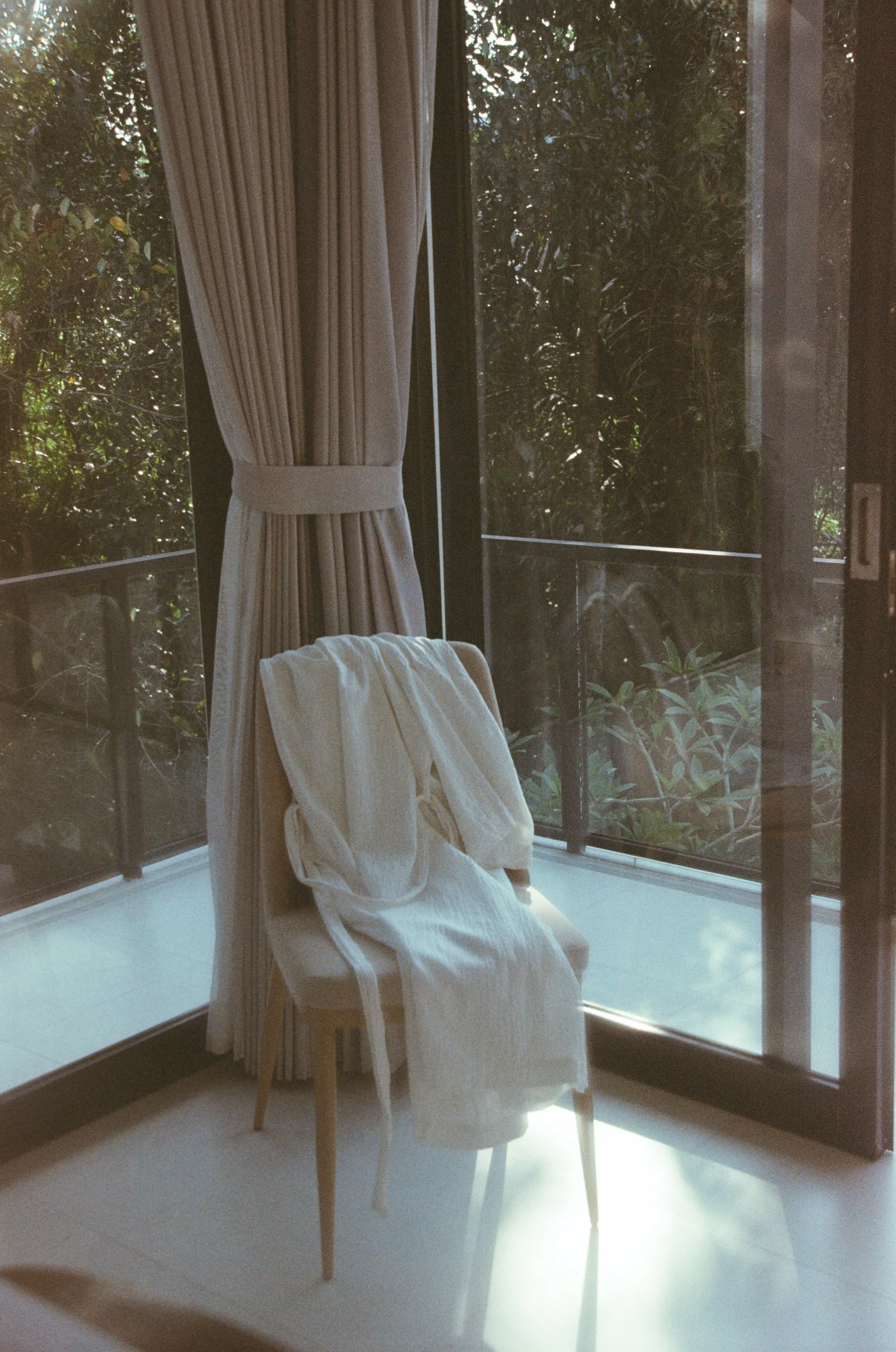 A chair with a white cloth draped over it, positioned in front of large glass sliding doors, with a view of green foliage outside, and beige curtains partially drawn to the side.