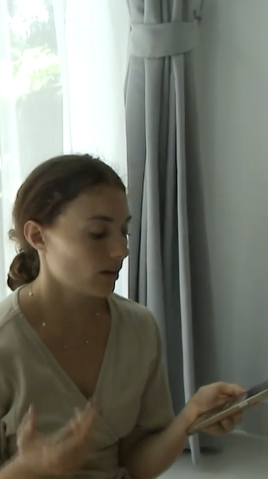 A woman, wearing a beige shirt, talking on the phone to someone receiving healing remotely, while sitting near a window.