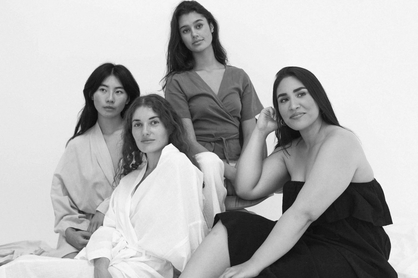 Four women sitting and standing together in a studio, dressed in different outfits, with a plain white background.
