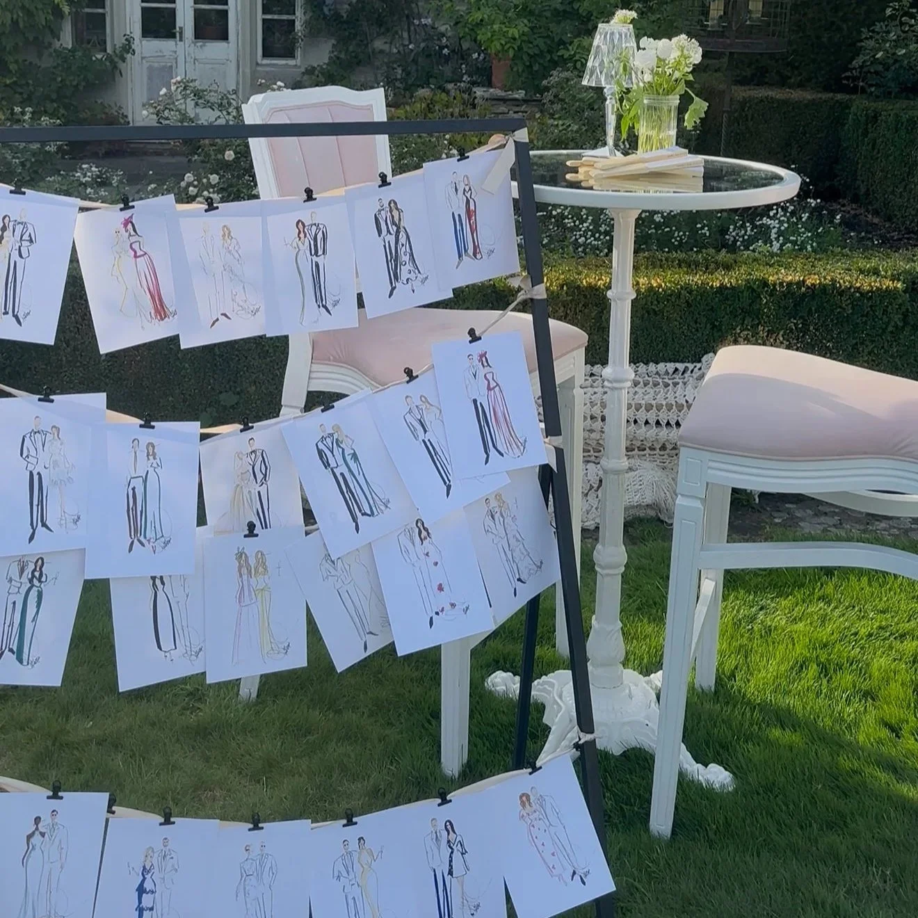 Outdoor garden scene with live fashion illustrations of guests hanging on a black display frame, a white round table with a flower vase, a pink upholstered chair, and green foliage in the background.