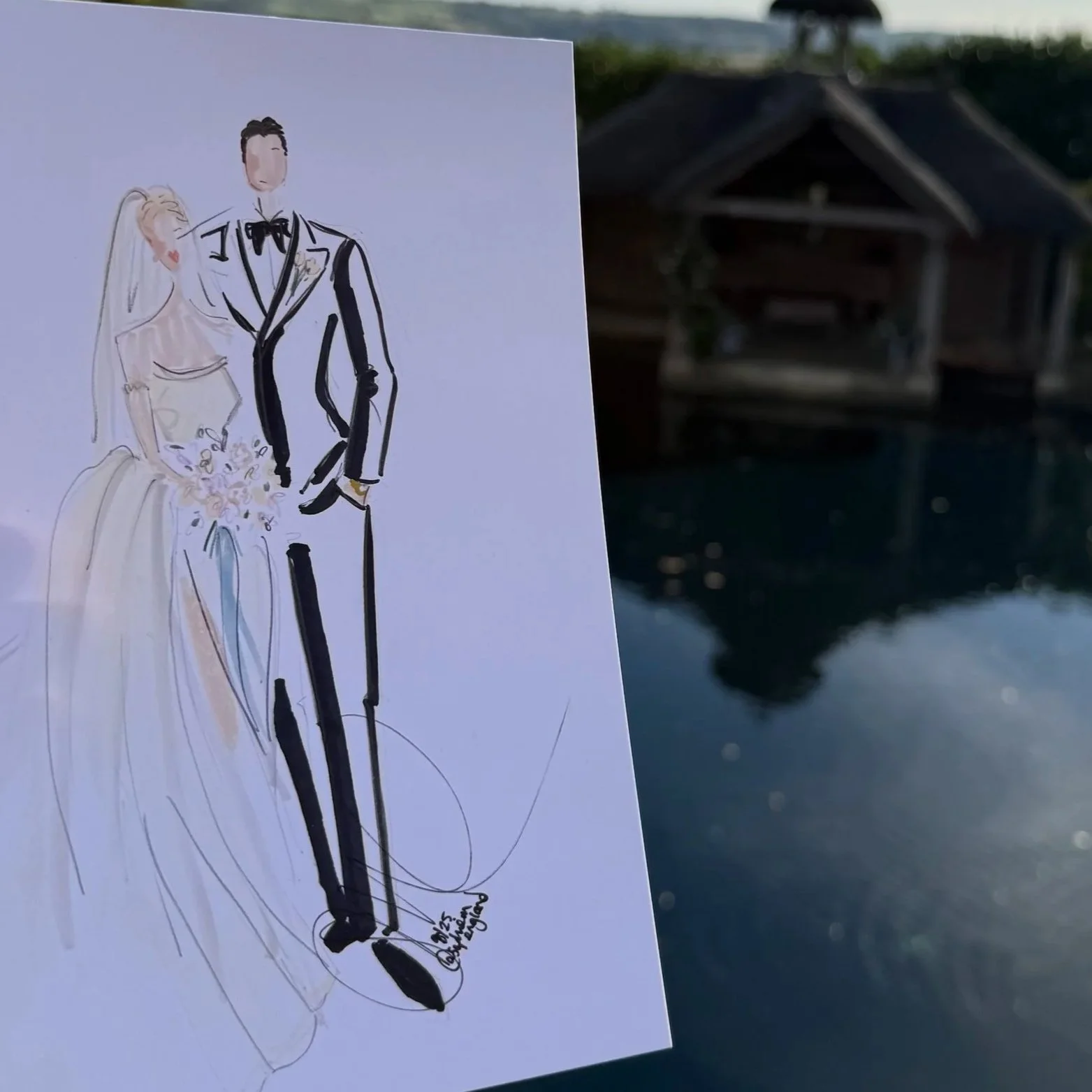 A hand holding a live art fashion drawing of a bride and groom in front of a pond with a wooden pavilion and trees, under a blue sky with some clouds.