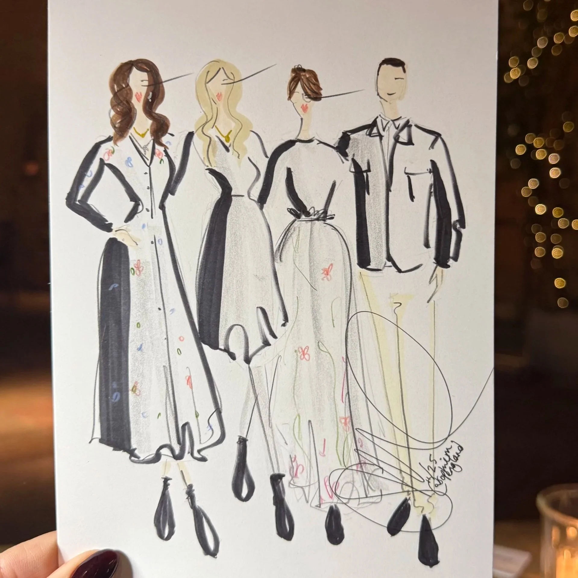 A hand holding a sketch of four fashion models in stylish outfits. The background shows a dimly lit indoor space with warm lighting, candles, and some writing tools on a table.