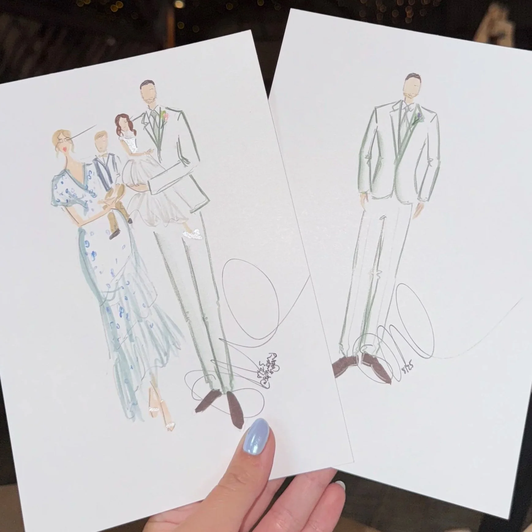 Hand holding two drawings of a wedding party, one colored and one outline, in front of a ceiling decorated with string lights.