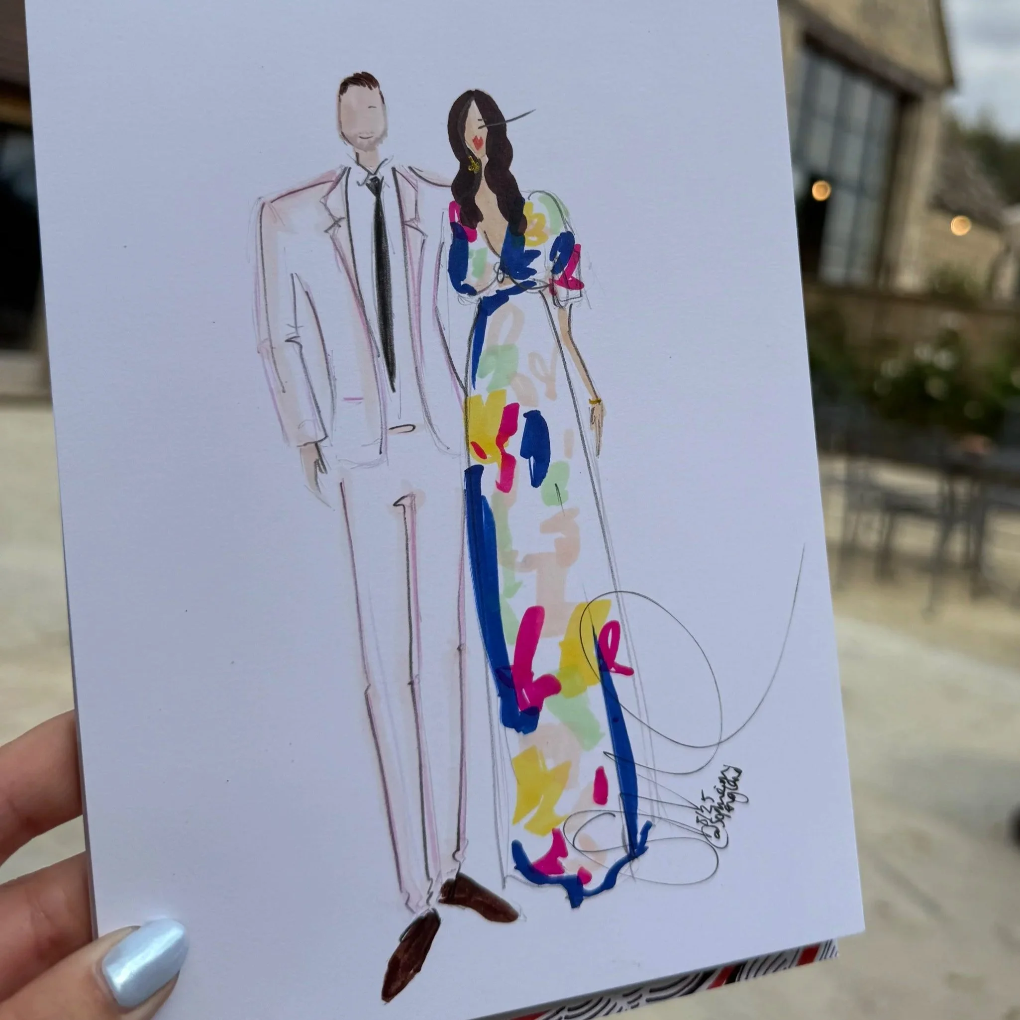 Hand holding a colorful illustration of a man in a suit and woman in a vibrant dress, with outdoor seating and a cloudy sky in the background.