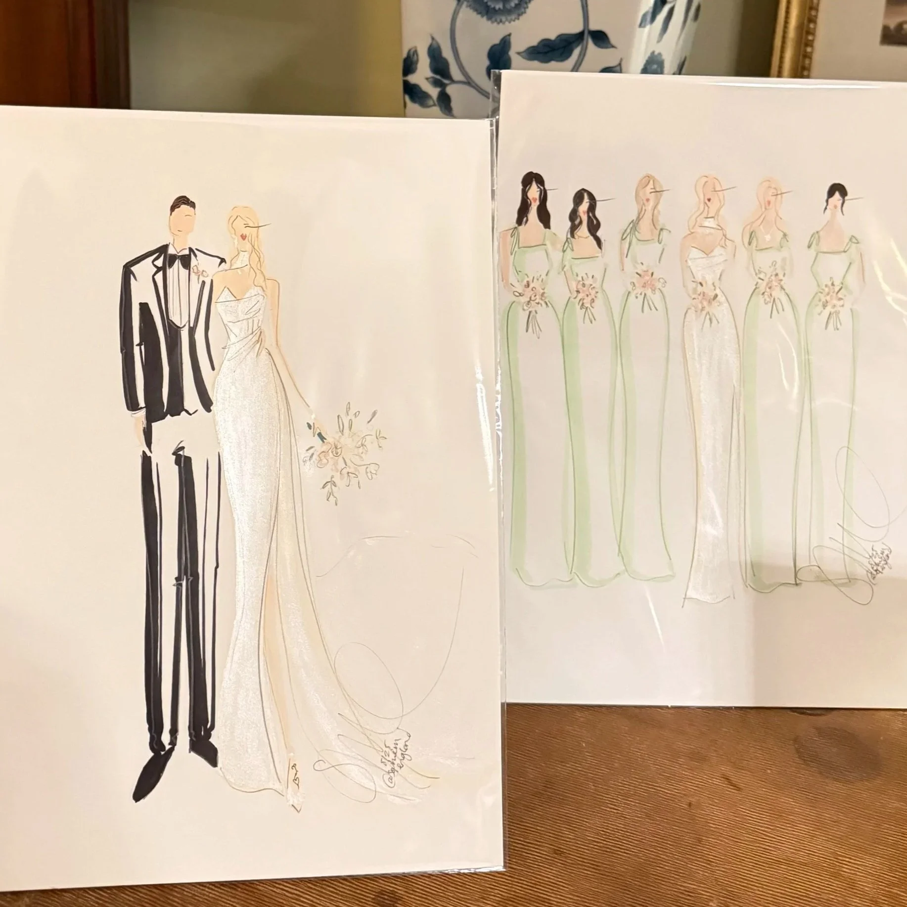 Two fashion sketches of wedding dresses and bridesmaids dresses placed on a wooden table.