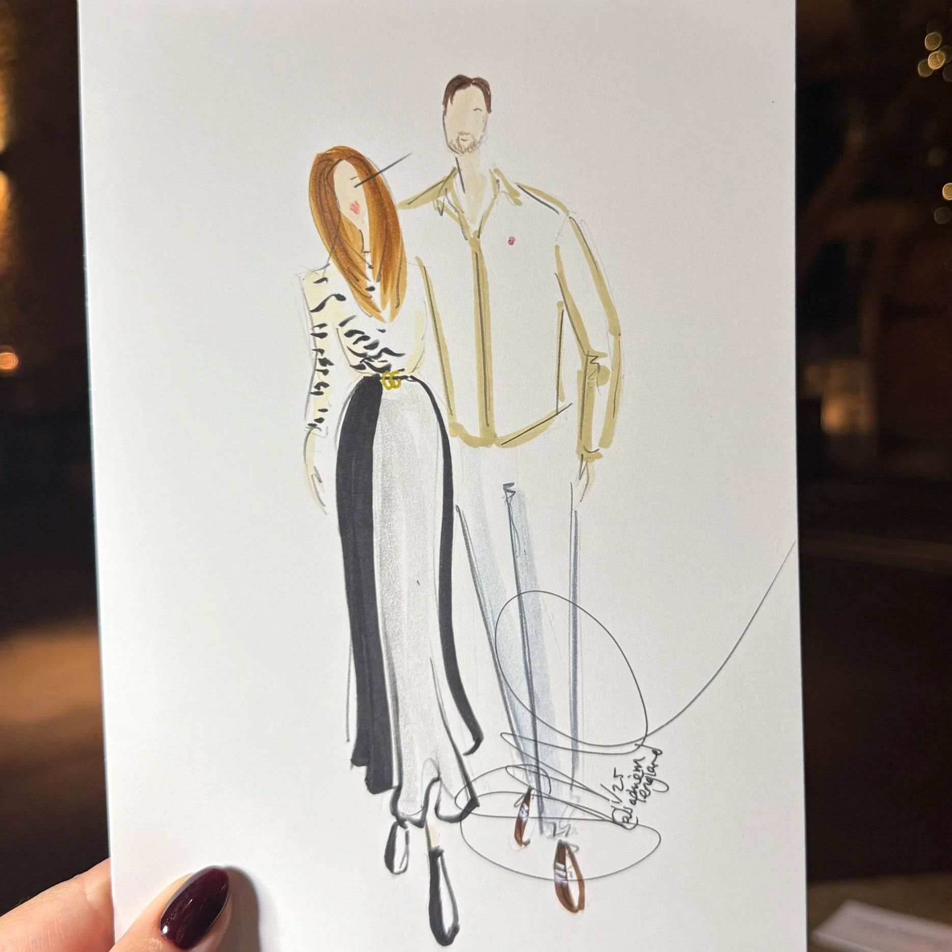 A hand holding a sketch of a man and woman in fashionable clothing, with a brick wall and string lights in the background.