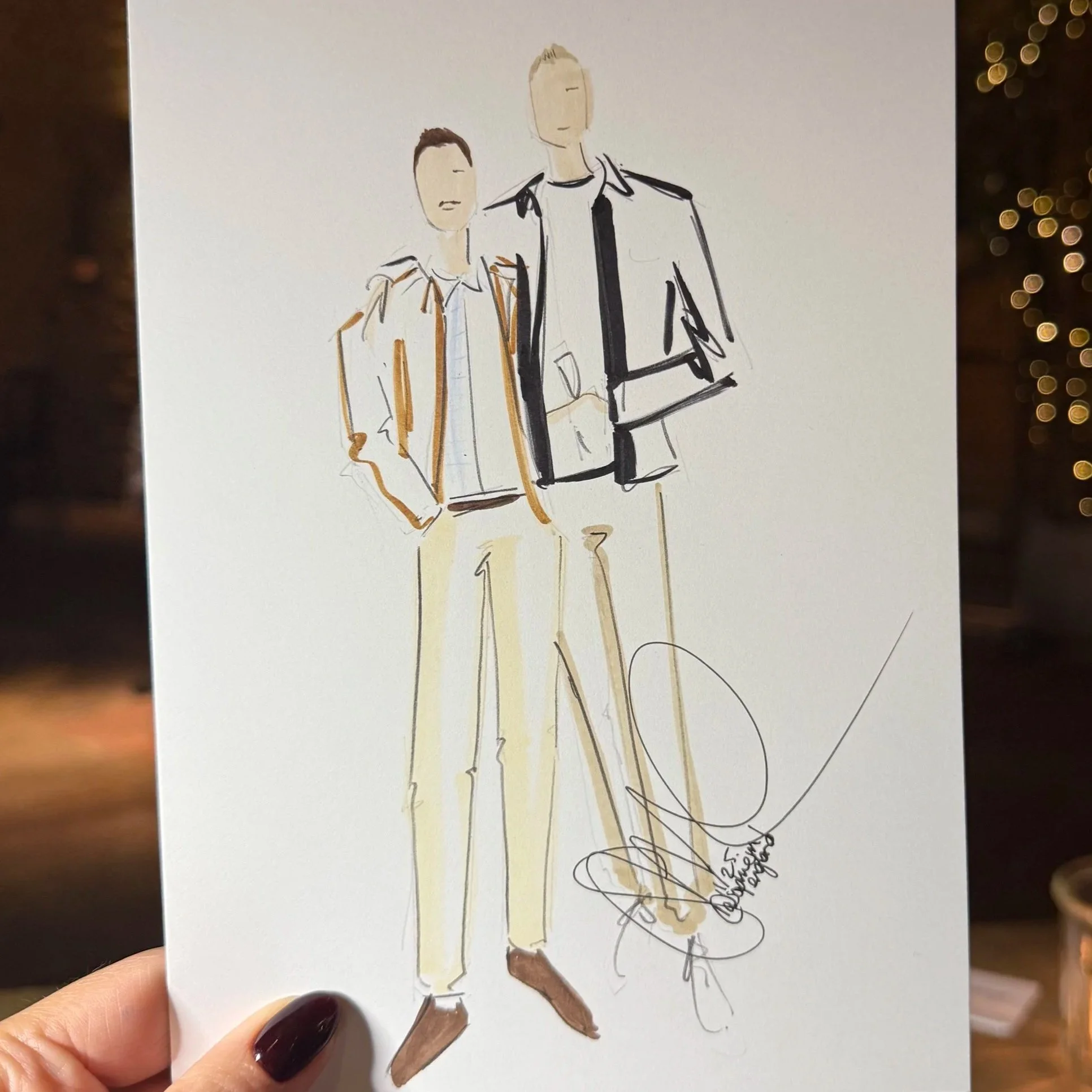 A hand holding a sketch of two men in fashionable clothing. The background is a dimly lit room with fairy lights and a candle on a wooden table, with various markers visible.