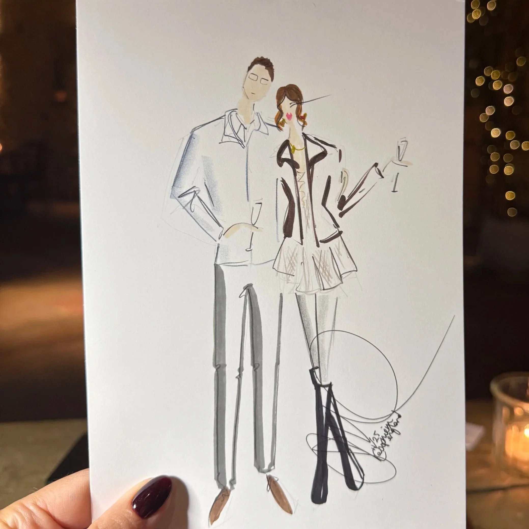 A hand holding a fashion sketch of a man and woman, with the man in a blazer and trousers and the woman in a skirt, blazer, high boots, and holding a glass of wine, against a warm, dimly lit background with fairy lights.