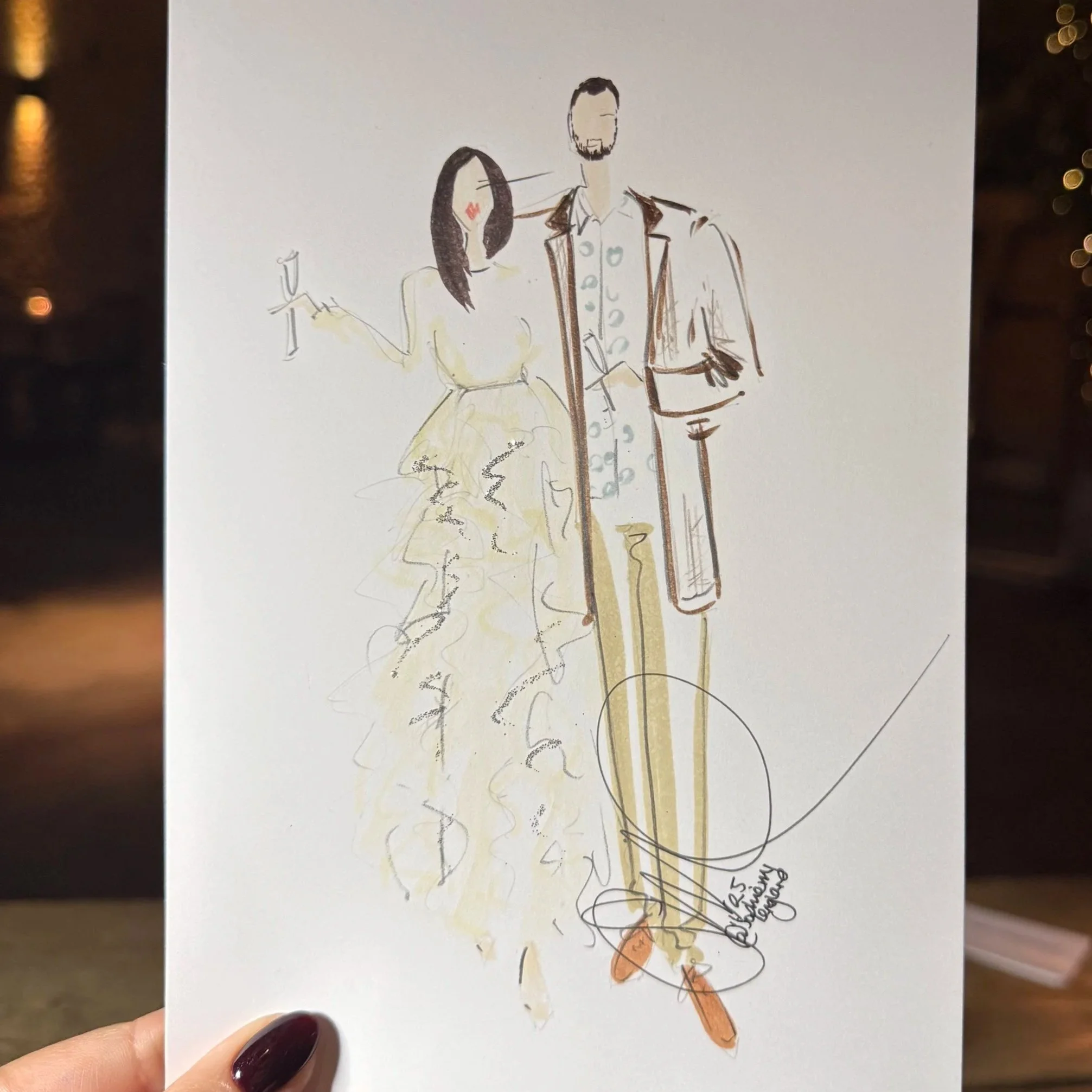 Hand-drawn live sketch of a couple, a woman in a flowing dress and a man in a suit, standing next to each other. The woman holds a glass of wine, and both figures are outlined with loose, artistic strokes. The sketch is held up in front of a warmly l