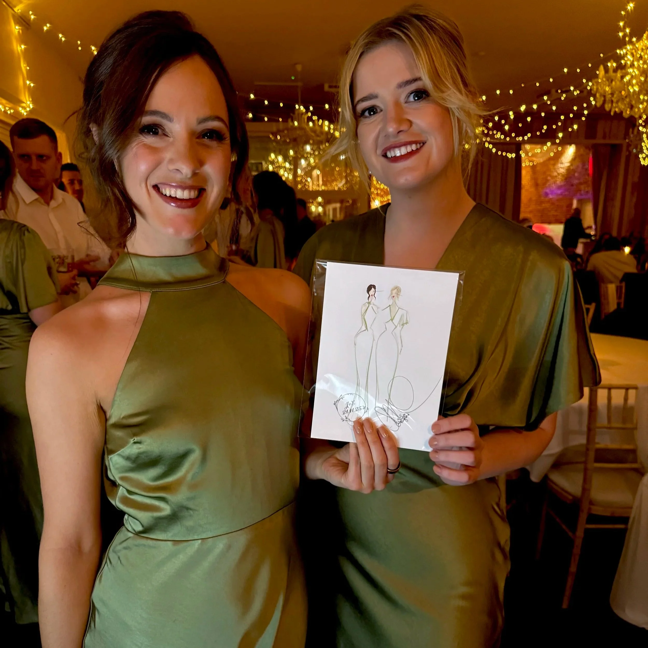 Two women in matching olive-green dresses smiling at a celebration or party, holding a drawing of fashion sketches, with warm lighting and festive decorations in the background.