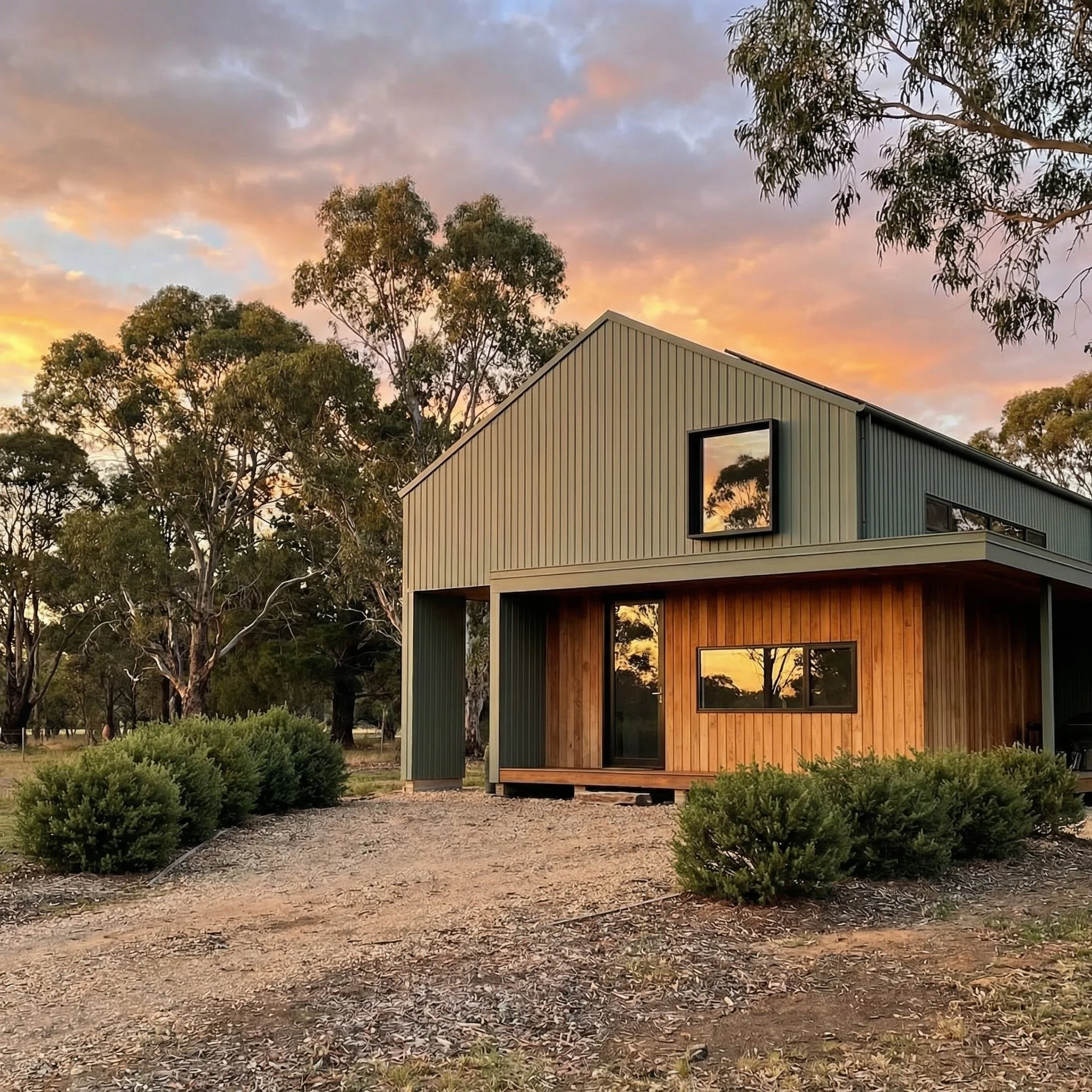 A modern house with vertical siding, large windows, and a metal roof, situated among trees during sunset with colorful clouds in the sky.