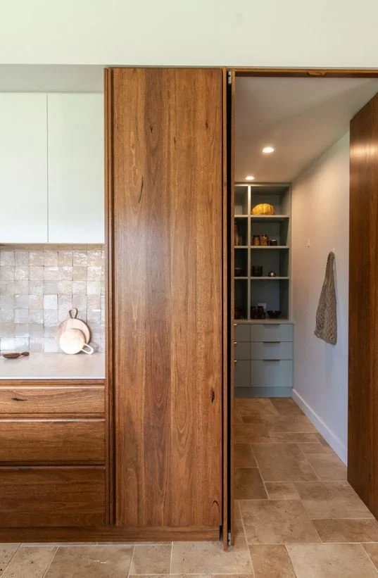 View of a kitchen with wooden and white cabinets, a door reveals a pantry with shelves and drawers, beige tiled floor, and a beige towel hanging on the wall.
