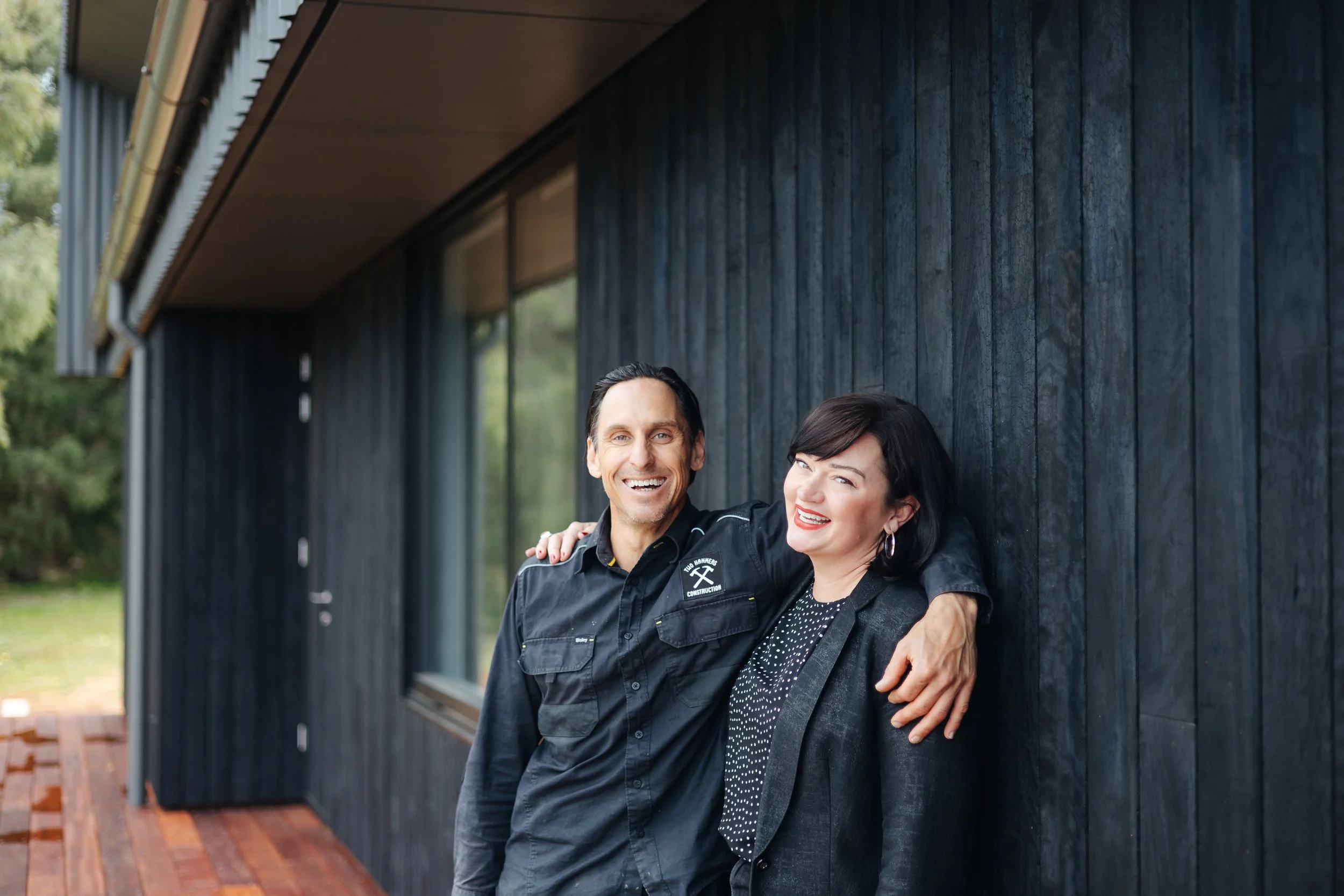 A man and woman smiling and hugging each other outside a modern house with dark wood siding and large windows.