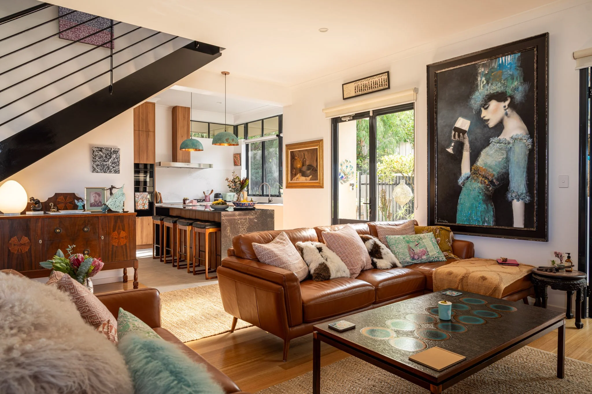 Interior of a living room with a leather sofa, pillows, a coffee table, and large wall art, kitchen and dining area in the background with pendant lights and windows showing greenery outside.