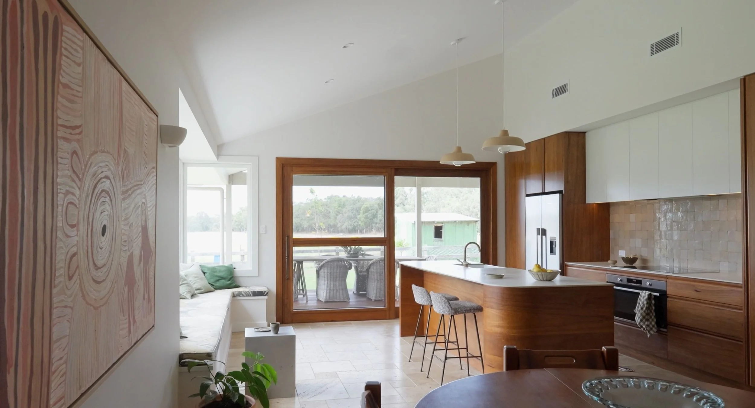Interior view of a modern kitchen and living area with natural light, wooden accents, a large window/door, a kitchen island with stools, and a glimpse of outdoor seating.