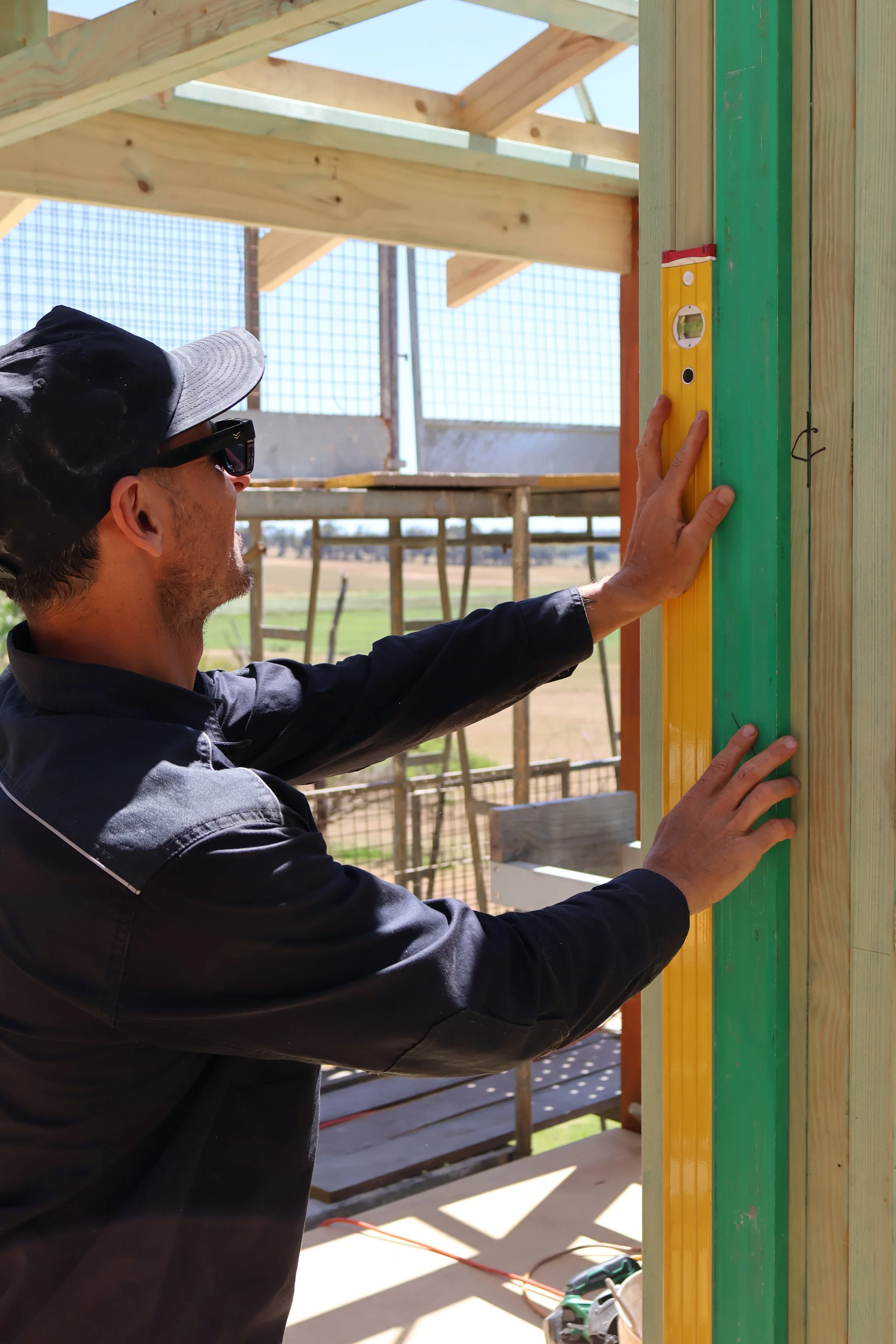 A man wearing a black jacket, a gray baseball cap, and safety glasses is measuring a wooden post with a yellow level at a construction site for a building. The background shows scaffolding, a wooden framework, and a rural landscape.