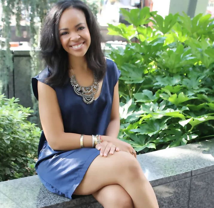 Smiling woman in a blue dress sitting outdoors on a stone ledge, surrounded by green foliage.
