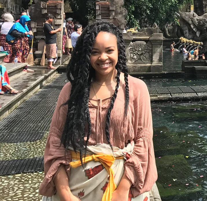 Smiling woman with long twisted hair standing near a water feature at a traditional Asian temple, with people in colorful clothing and temple architecture in the background.