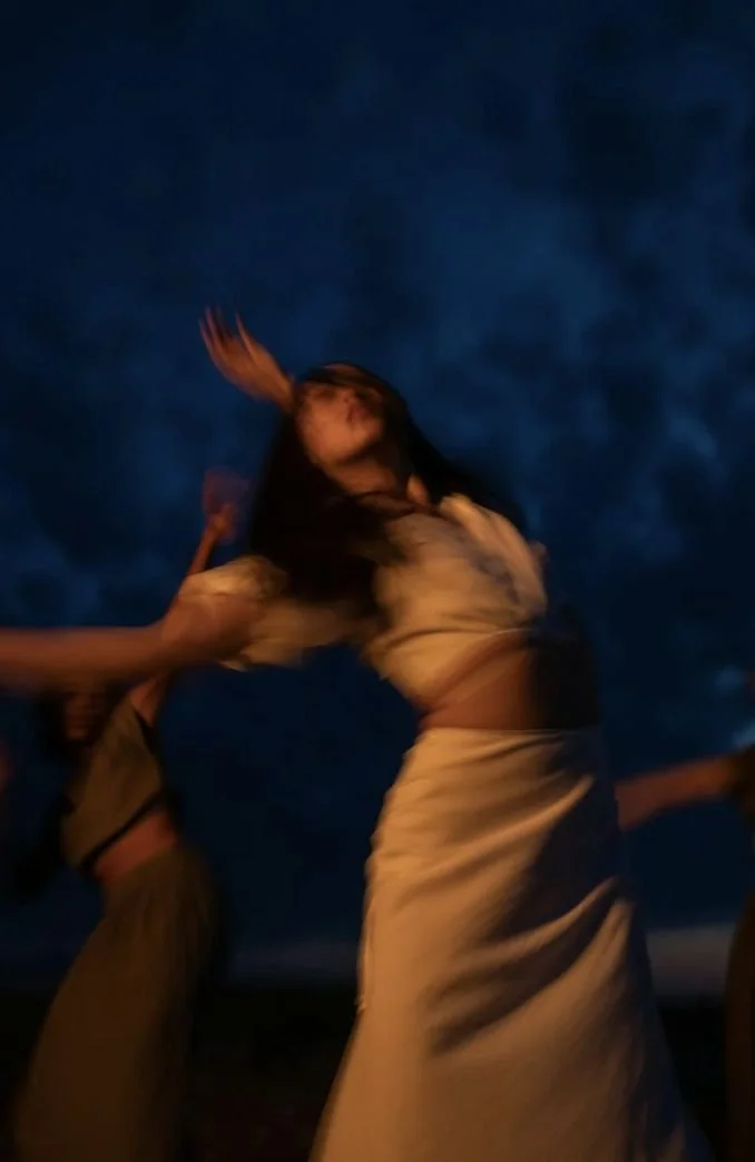 A woman is dancing outdoors during dusk or night, with a partly cloudy dark blue sky in the background.