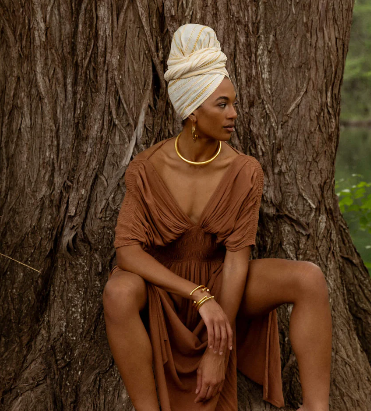 A woman sits against a large tree trunk, wearing a brown dress and a white head wrap, accessorized with gold jewelry, looking to the side.