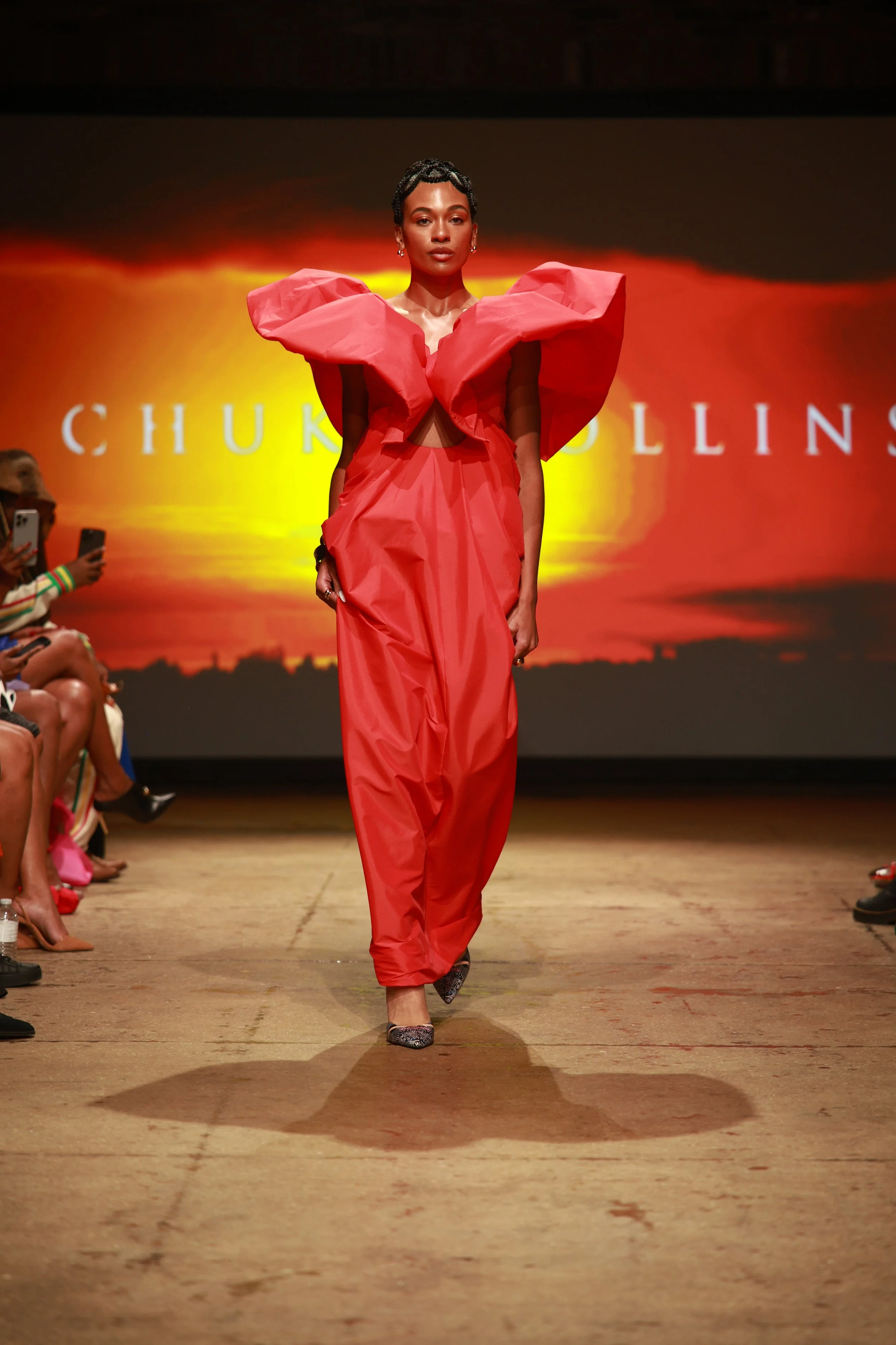 A model walking on a runway during a fashion show, wearing a red dress with large ruffled shoulder details and high heels, with an audience watching.