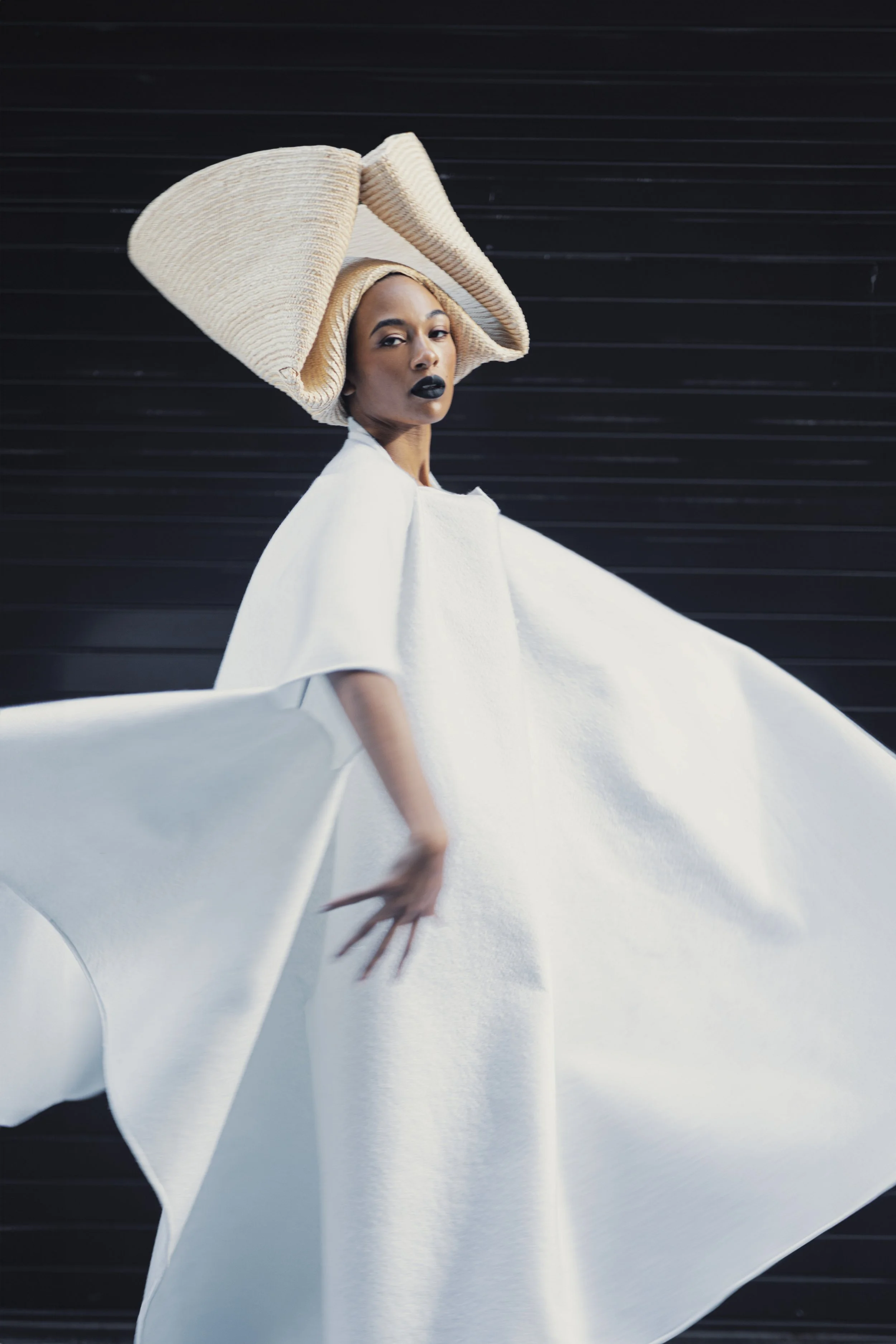 A woman wearing a large, wide-brimmed, woven hat and black lipstick posing against a dark, striped background, with an elegant white flowing garment.