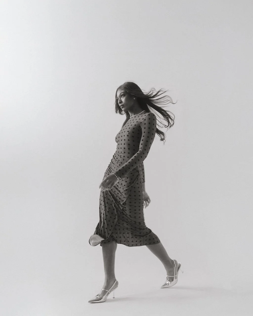 A black and white photo of a woman in a polka dot dress with flowing hair, walking in a studio with a plain background.