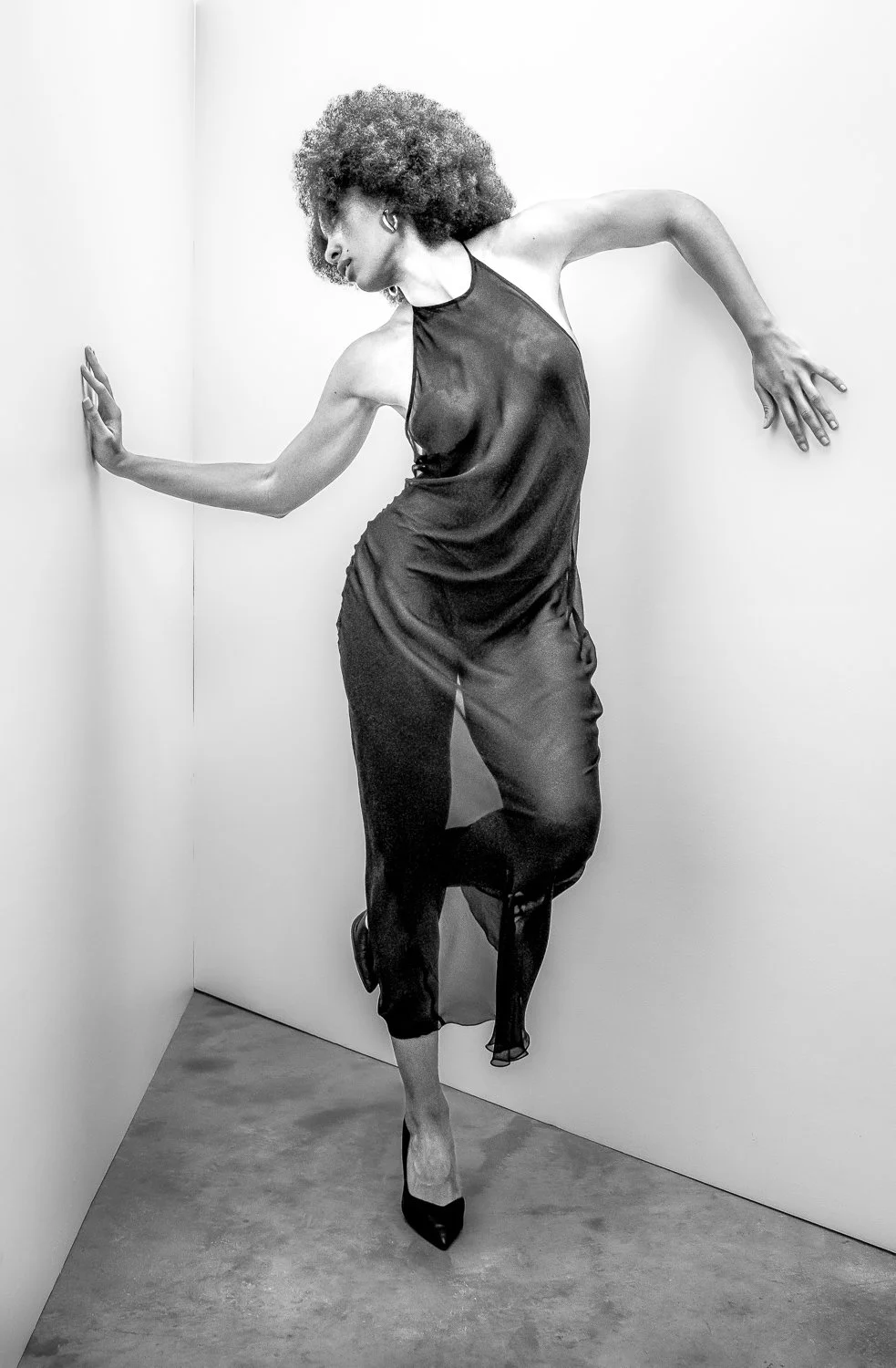 A woman with curly hair in a black dress and high heels striking a pose with her hands against the wall in a corner.