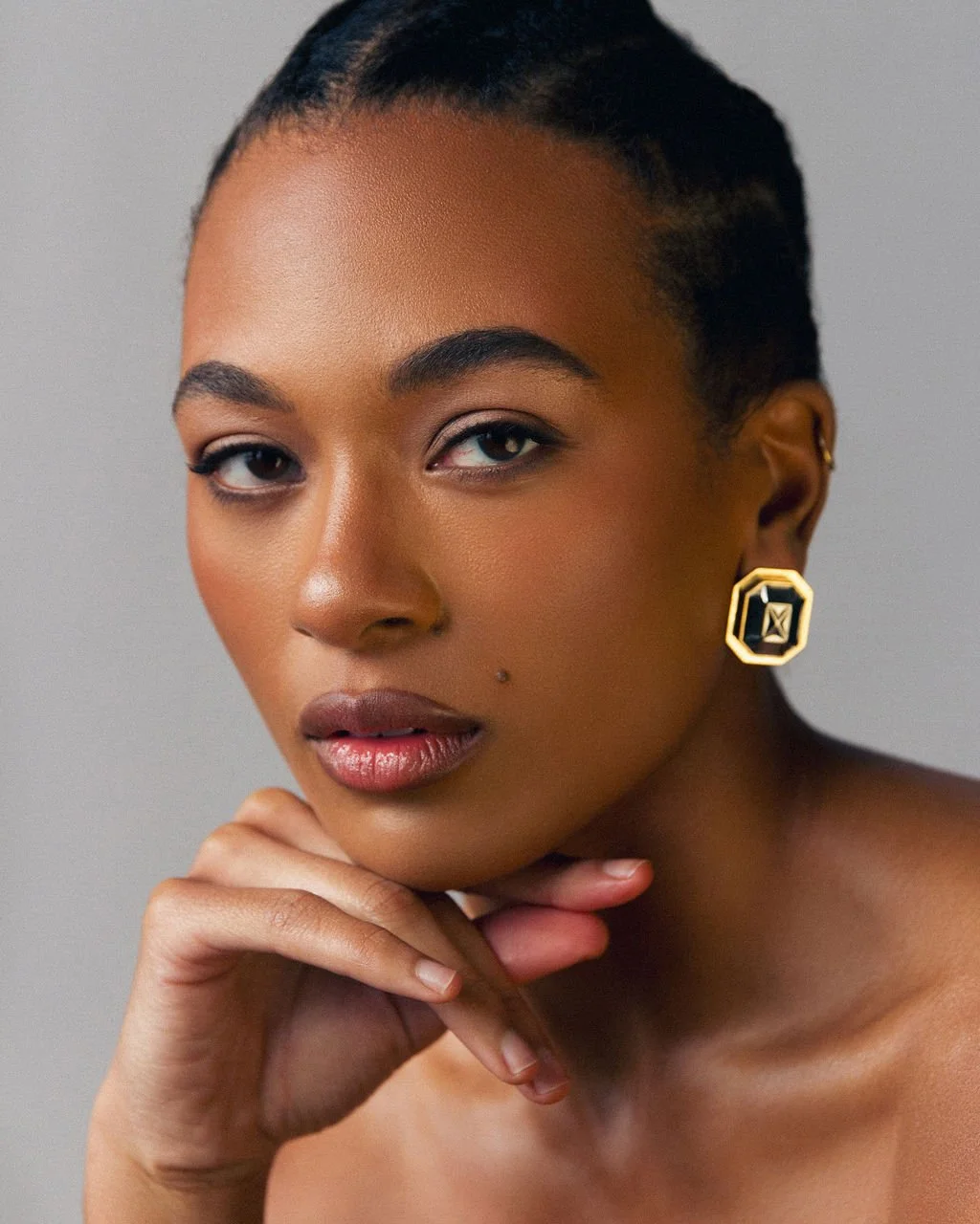 Close-up portrait of a woman with a confident expression, short dark hair, perfect makeup, and wearing statement gold earrings.
