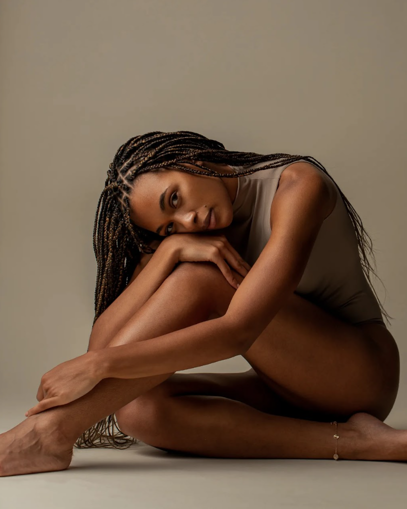 A woman with long braided hair sitting on the floor, resting her head on her knee, wearing a beige sleeveless top and a delicate ankle bracelet.