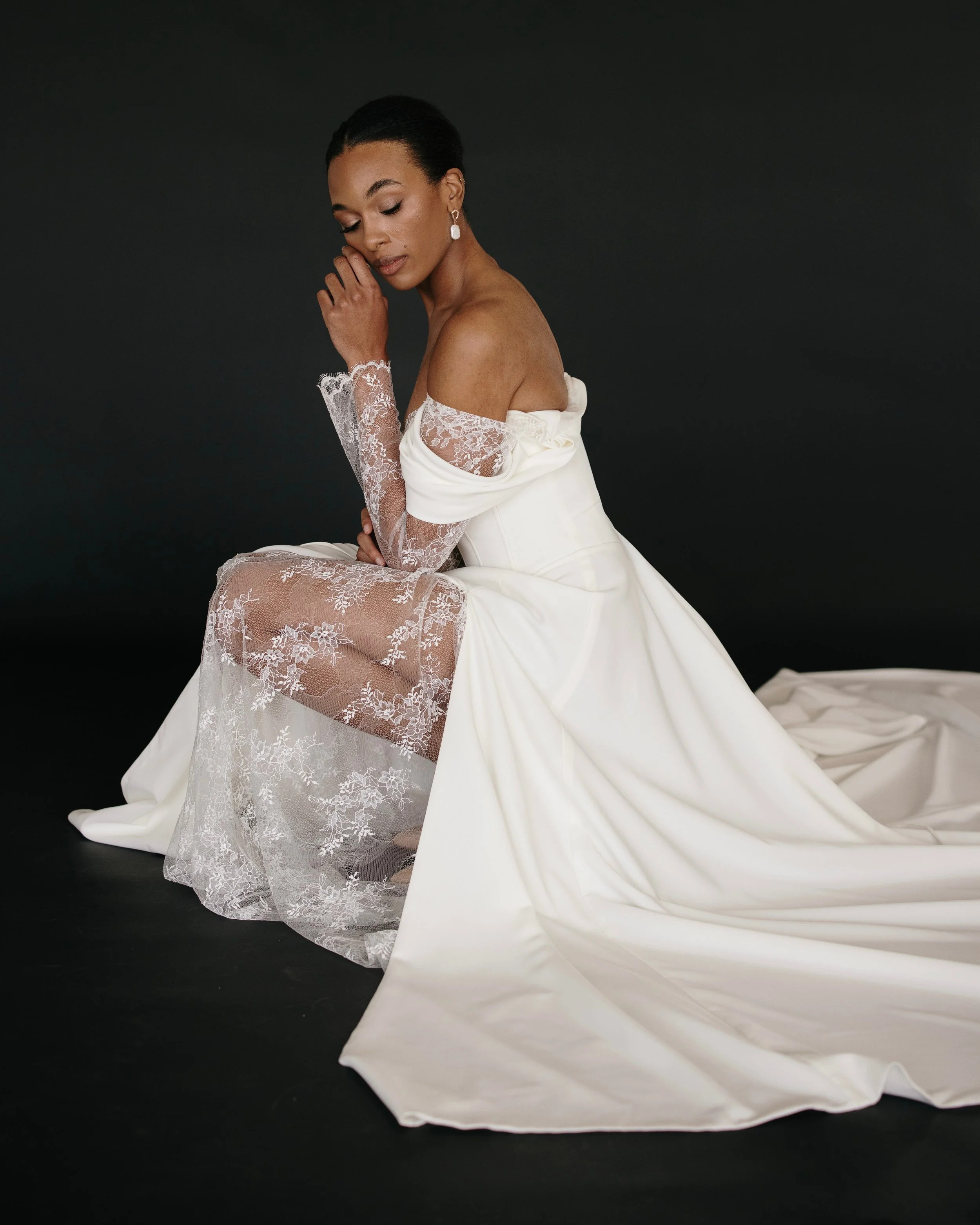 A woman with dark hair styled back, wearing a white dress with off-the-shoulder lace sleeves, sitting against a black background, with her eyes closed and her hand resting near her face.
