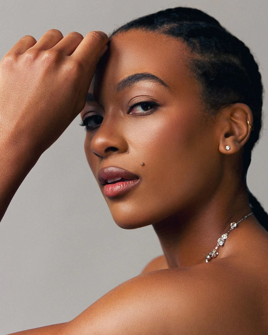 Close-up of a woman with short braided hair and medium skin tone, looking at the camera with her hand resting on her forehead. She has pierced ears with diamond studs, a nose piercing, and is wearing a delicate necklace.