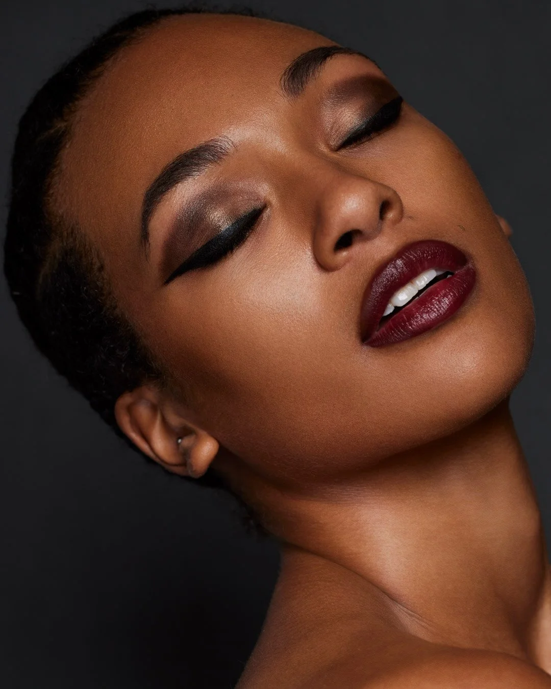 Close-up of a woman with dark makeup, closed eyes, and deep red lipstick, with a dark background.
