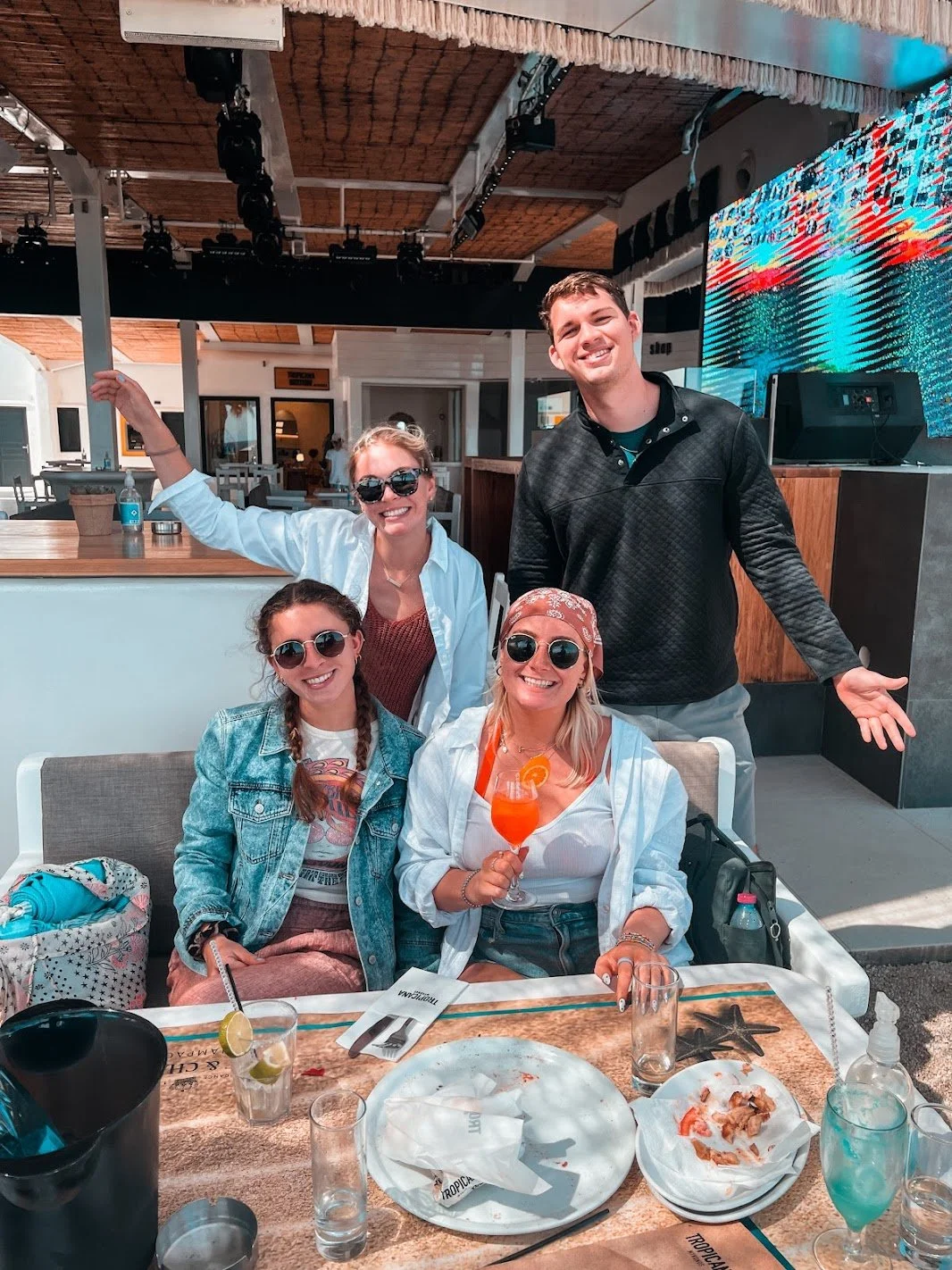 Four partiers pose with drinks in front of a DJ booth at Tropicana Day Club in Mykonos.