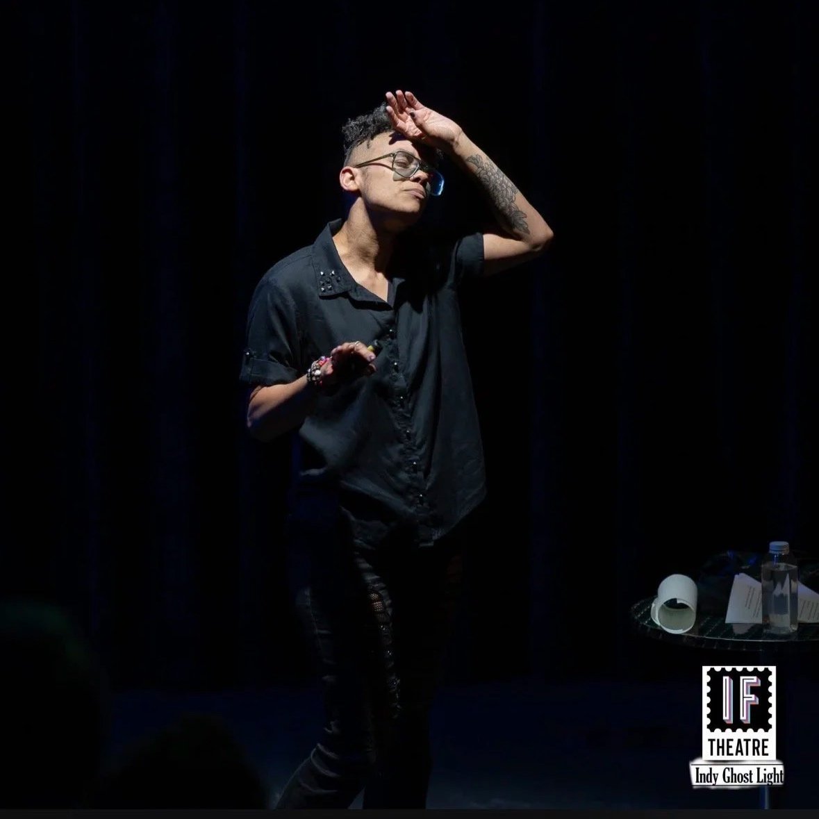 Photo of Carrie dressed in all black in black space with arm over their forehead looking forelorn