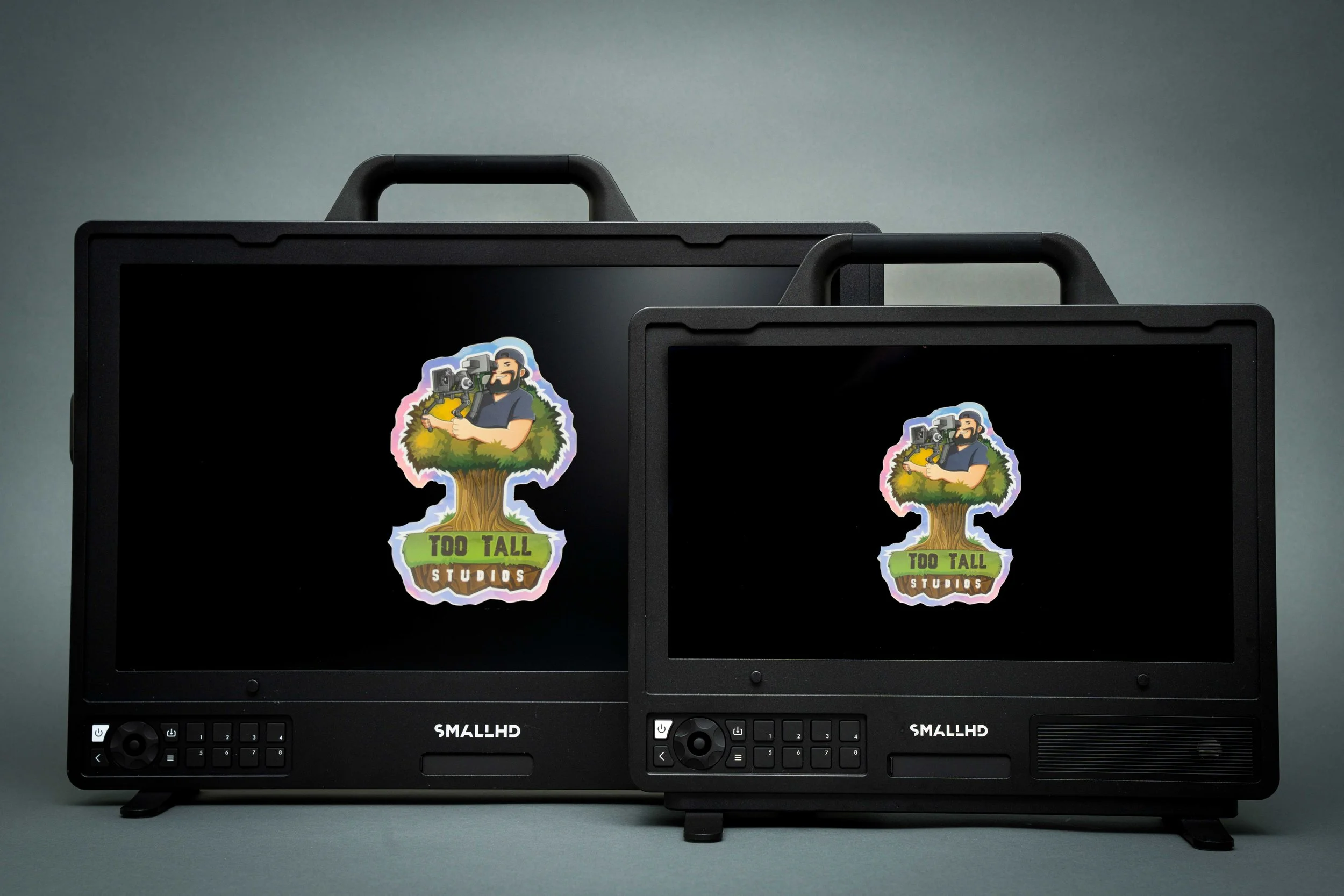 Two small portable monitors with stickers on the screens. The stickers feature a cartoon man with a beard holding a camera, sitting on a tree, with the words 'Too Tall Studios'. The monitors are black, with controls on the front, and are placed against a plain background.