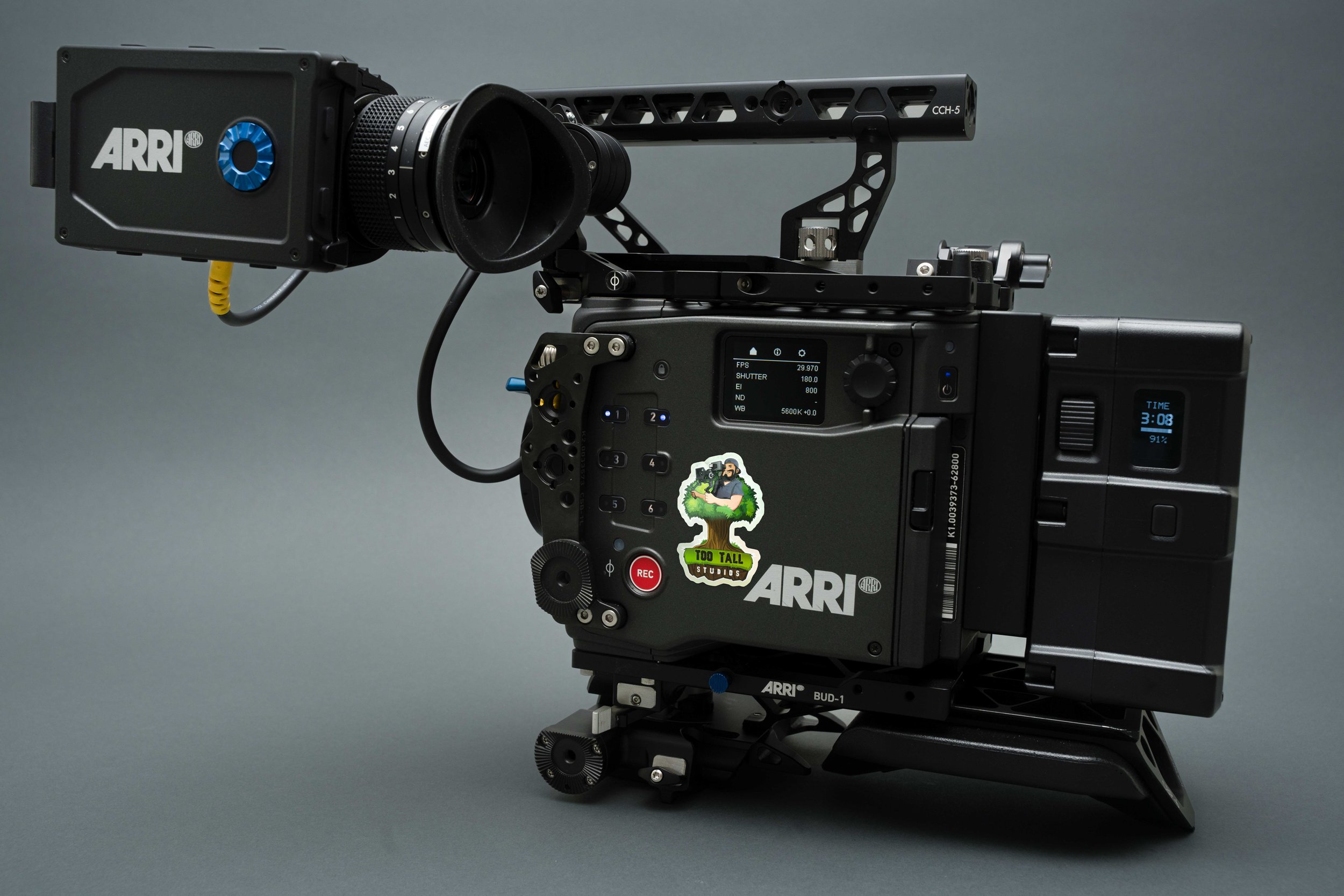 Professional cinema camera with matte box, mounted on a tripod, with ARRI branding and a sticker of a person holding a camera and the text "Too Tall Studios".