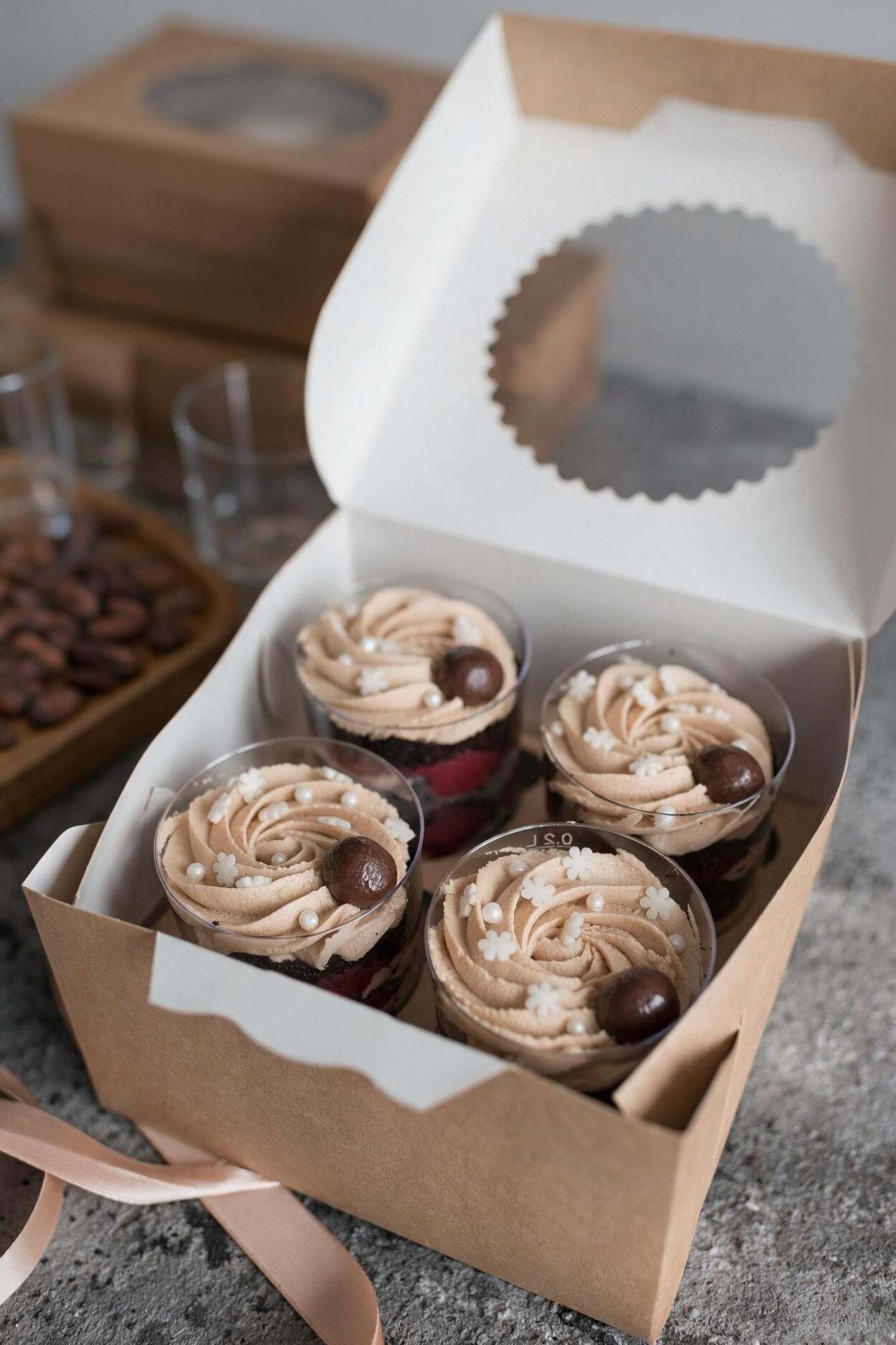 Four coffee-flavored cupcakes in clear cups with piped frosting, chocolate decorations and white sprinkles inside a bakery box.