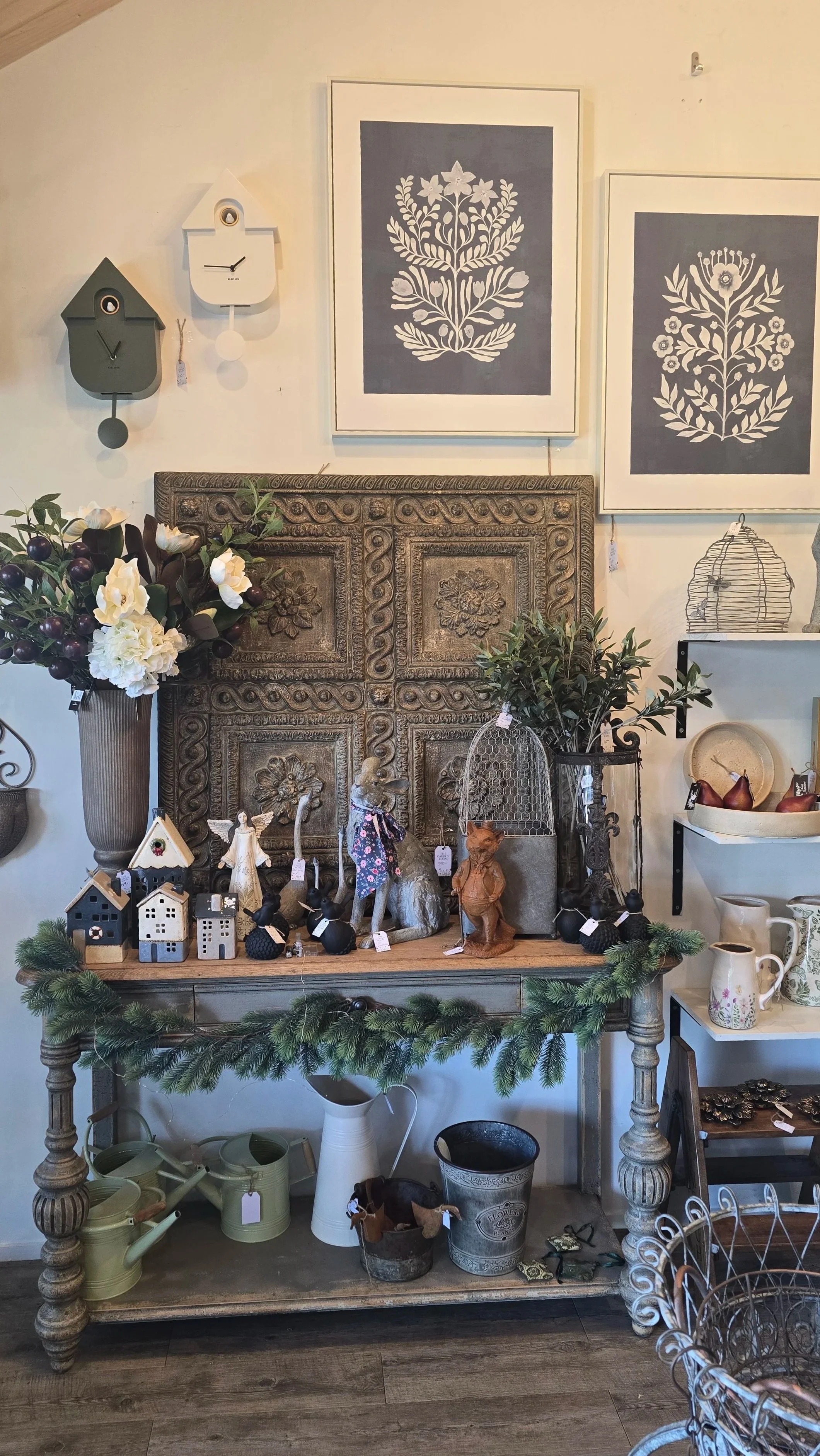 Decorative display of vintage and rustic items including a large carved wooden panel, framed botanical prints, ceramic and metal vases with flowers, small model houses, figurines, birdcages, and greenery on a wooden table with additional shelves hold