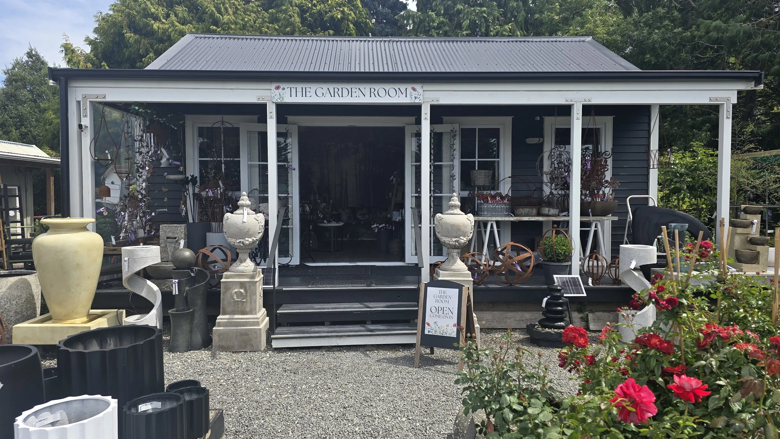 Exterior of a garden shop called The Garden Room with gardening decor and ornaments displayed outside. Sign indicates the shop is open.