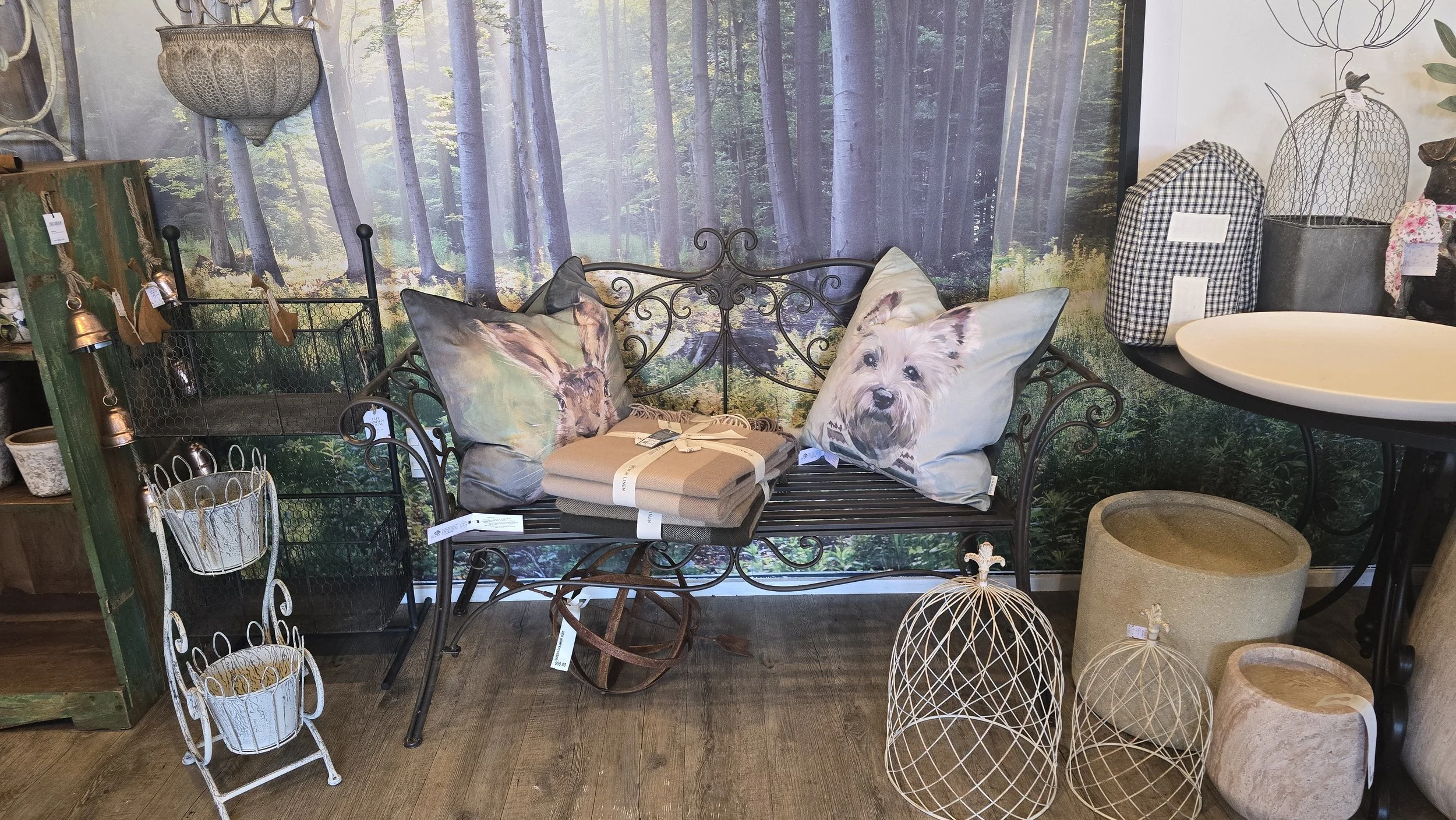 A display of decorative outdoor furniture and accessories in a store, featuring a garden backdrop with trees. There is a black metal bench with two pillows with dog images, a set of folded blankets, a tall white metal tiered plant stand, wire lantern