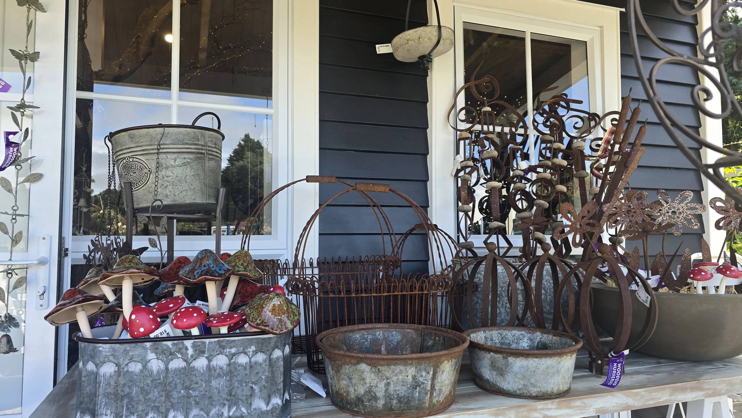 Display of gardening decorations including rusted metal plants, mushrooms, and baskets, outside a house with blue siding and a white-framed window.