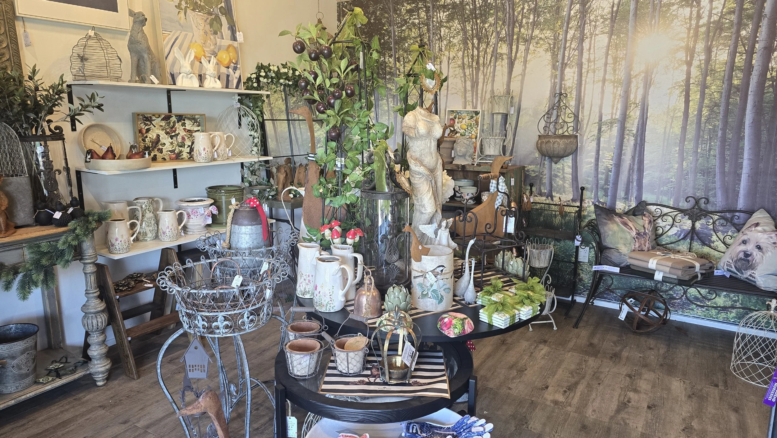 Interior of a gift shop displaying various decorative items including ceramics, metal figurines, plants, and pillows, with a forest scene mural background.