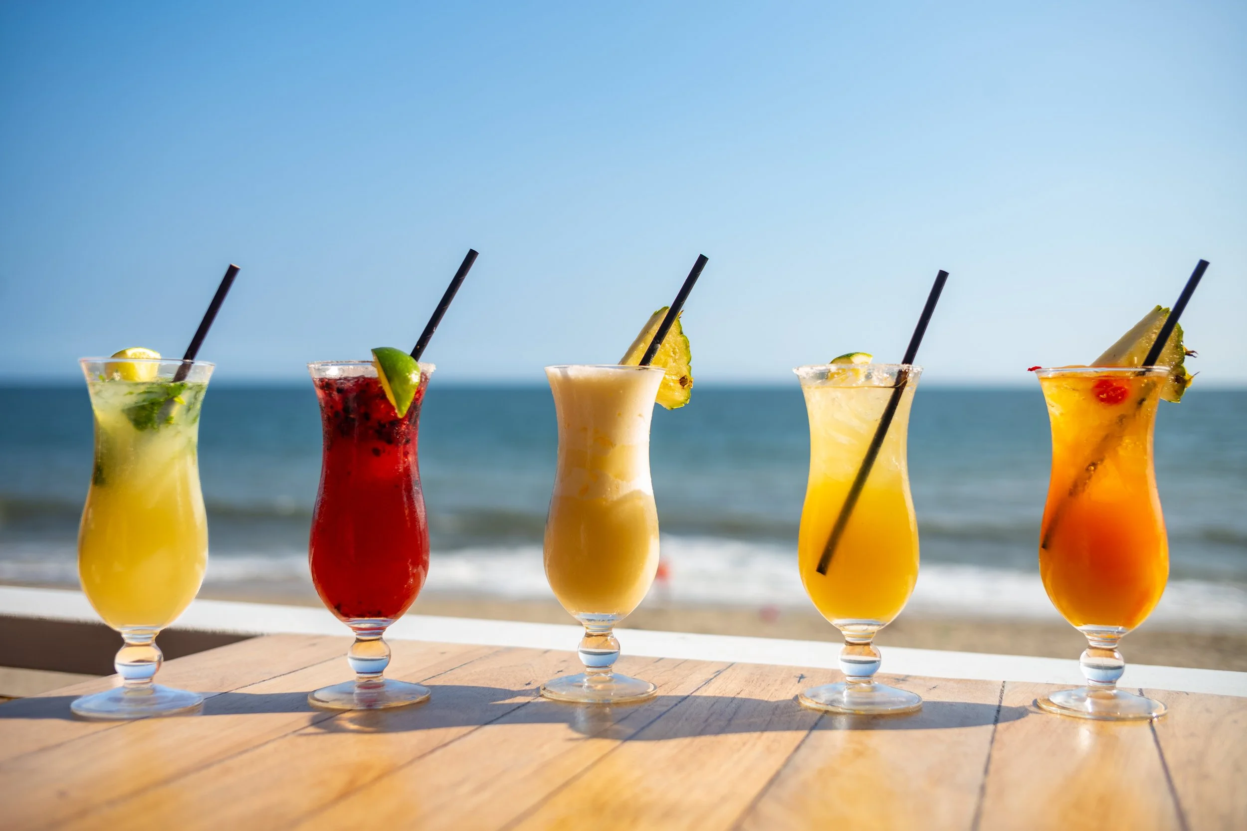 Five colorful tropical cocktails in tall glasses with fruit garnishes, placed on a wooden surface with a beach and ocean in the background.