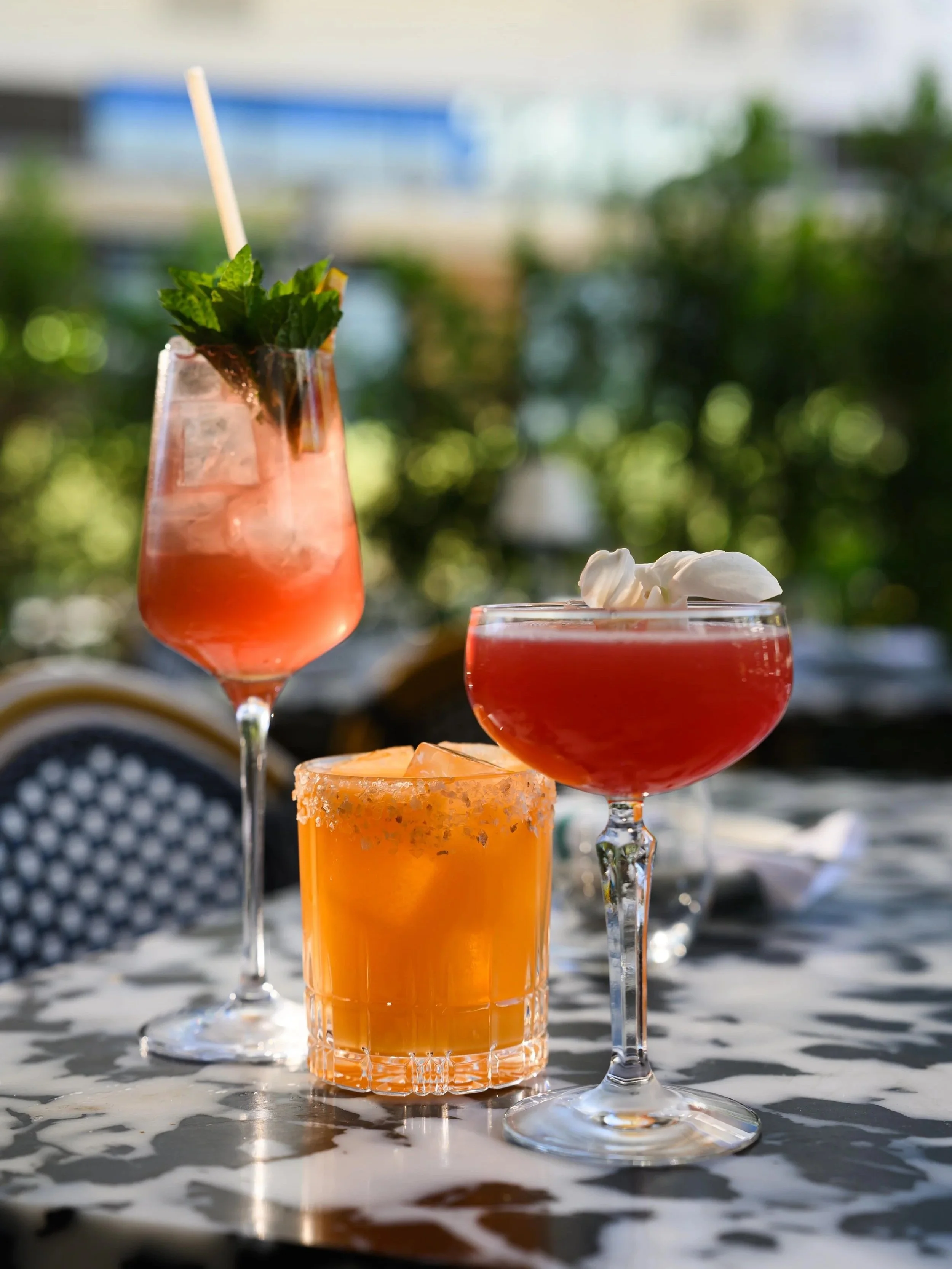 Three colorful cocktails on a table with a blurred green outdoor background. The drinks are garnished with mint and whipped cream, served in highball, coupe, and rocks glasses.