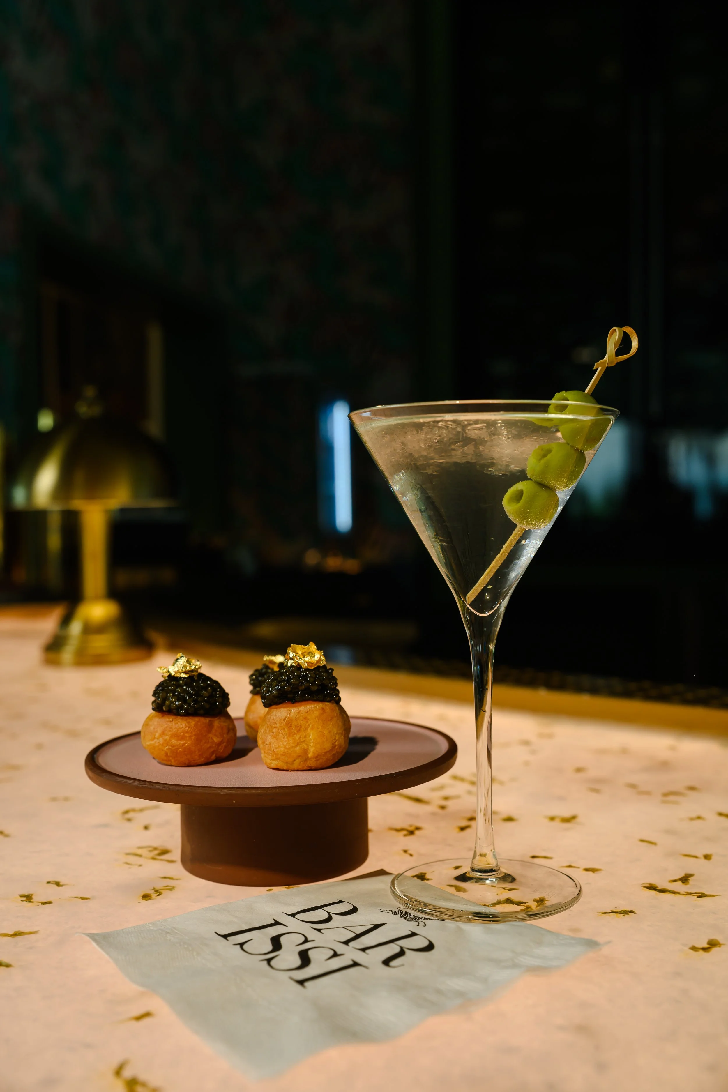 A martini glass with a green olive on a skewer, and three small pastries topped with black caviar and gold leaf on a pink and brown plate, set on a table with a decorative napkin reading "BAR LIST".